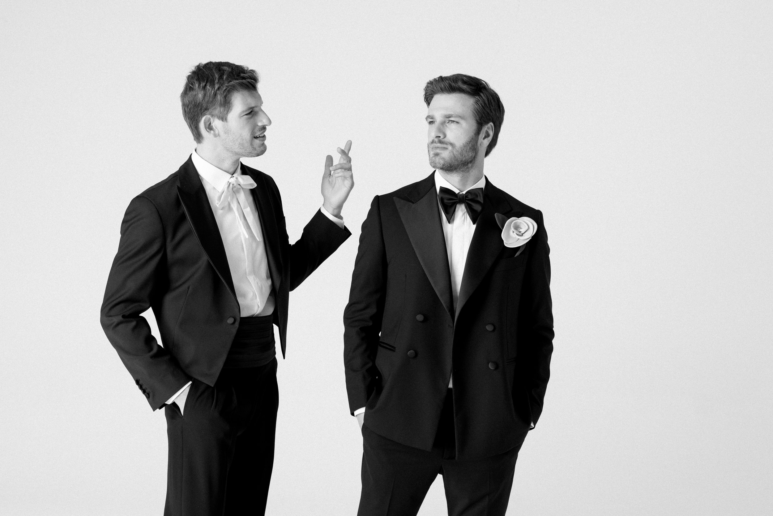 Due uomini in abiti eleganti discutono, uno con un gesto con la mano, l'altro con un'espressione seria, entrambi vestiti con abiti da sera con fiocco e fiore sulla giacca.