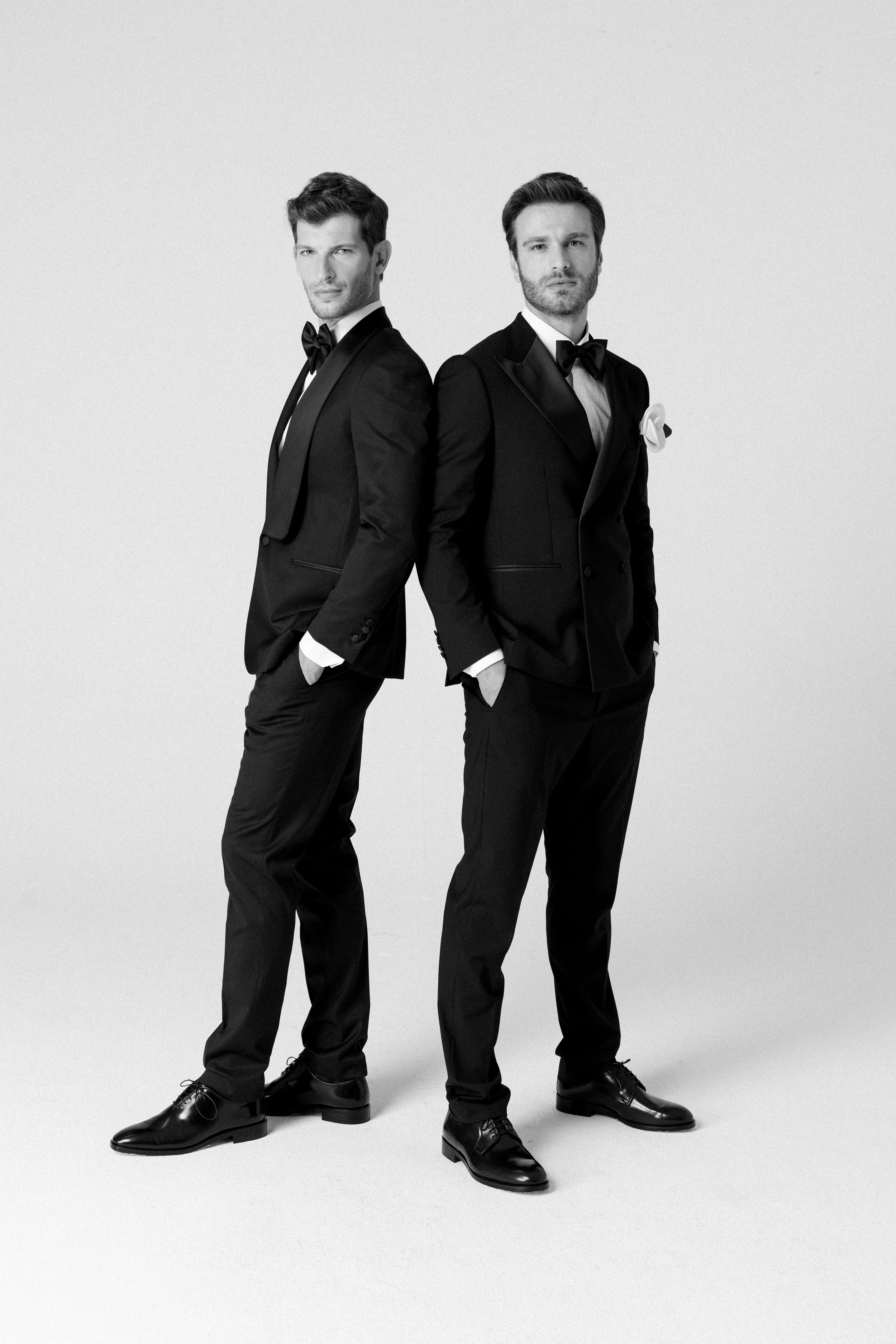 Due uomini eleganti vestiti con abiti neri e cravatte, uno con una rosa bianca in tasca, posano contro uno sfondo bianco in stile fotografico in bianco e nero.