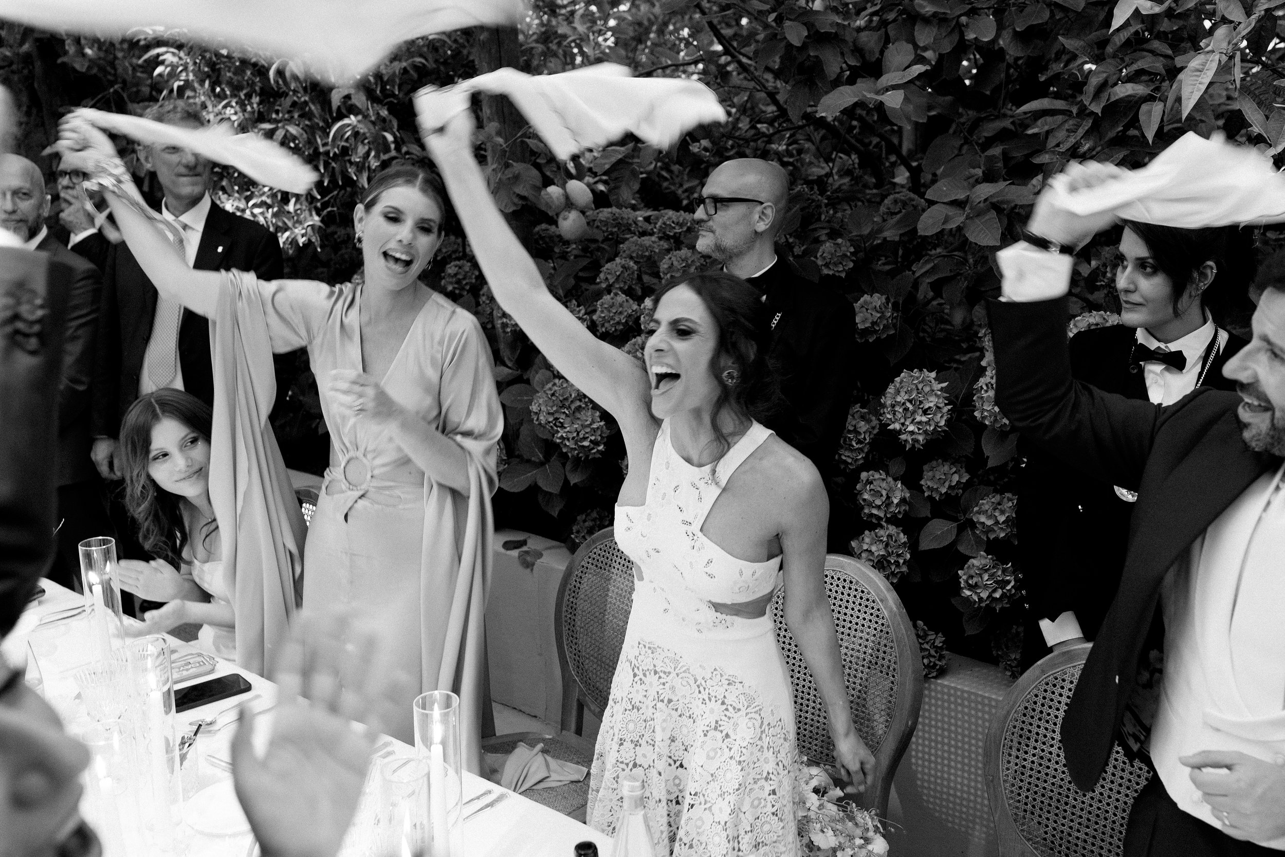 Un gruppo di persone scherza e si diverte a un ricevimento elegante, alcune fanno il tifo, ci sono fiori e bicchieri sul tavolo.