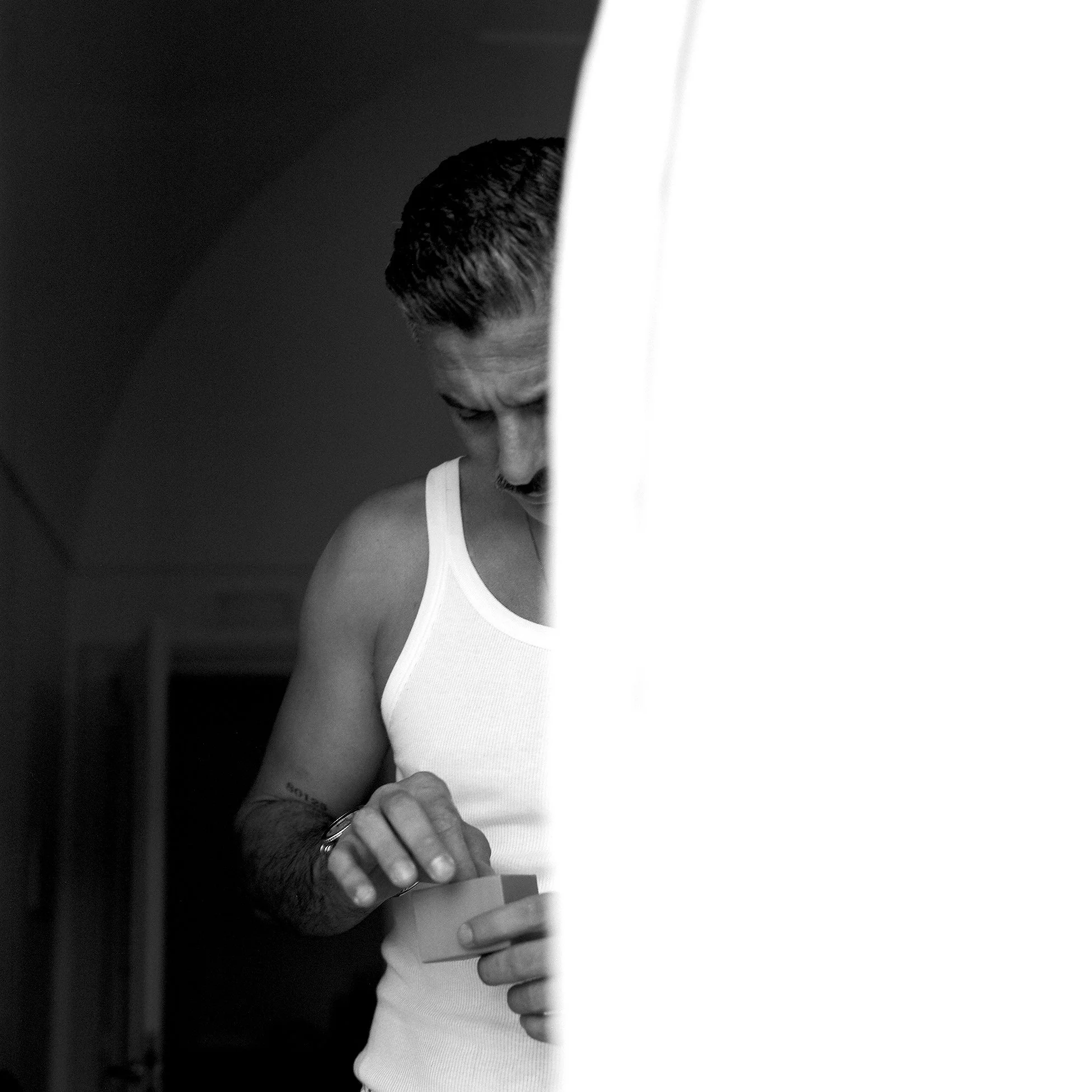 Un uomo con capelli corti, maglietta bianca, che si sta preparando, probabilmente per una performance o un evento, con un lucchetto al polso e un tatuaggio sulla spalla, sfila dietro una cortina.