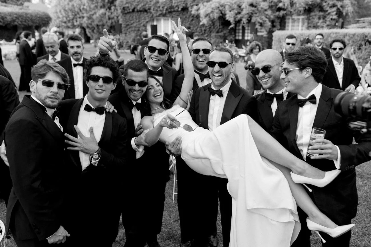 Un gruppo di persone vestite eleganti con completi neri e smoking, alcuni con occhiali da sole, che sollevano una donna in abito da sposa in modo giocoso durante un matrimonio all'aperto.