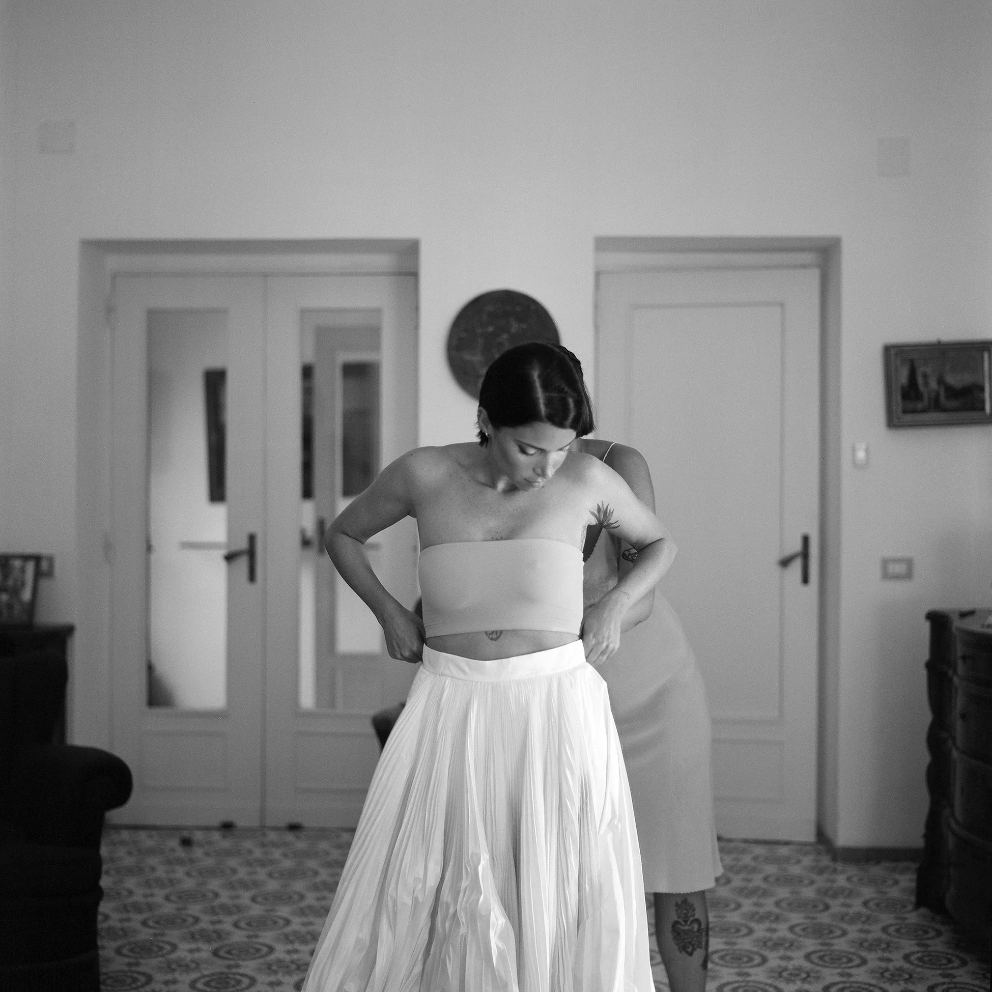 Una donna con capelli corti e tatuaggi, indossa un top senza maniche e una gonna lunga e ampia, si sta sistemando il vestito in una stanza elegante e ben arredata.