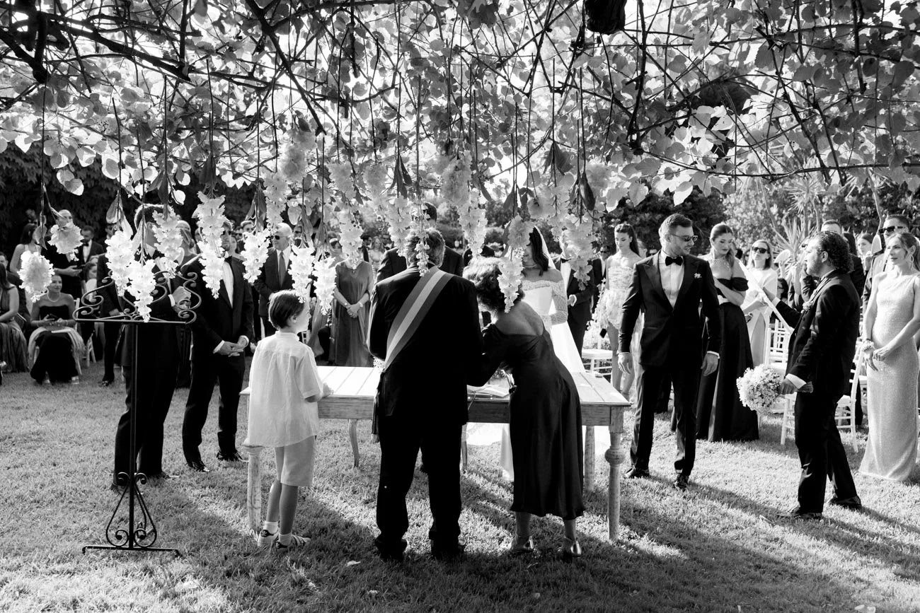 Cerimonia di matrimonio all'aperto con persone vestite elegantemente sotto un albero con fiori, in bianco e nero.