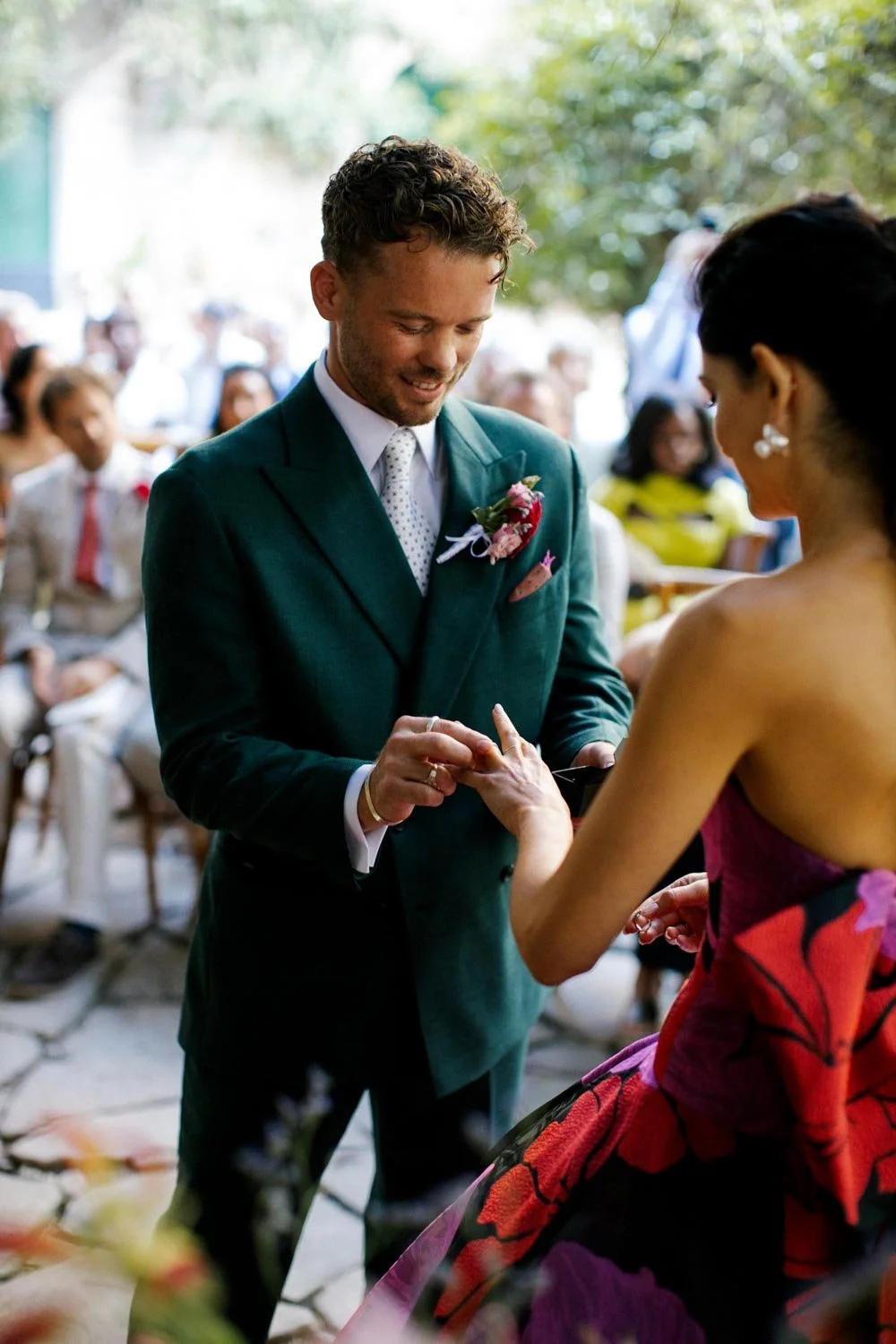 Uomo che scambia anelli con donna durante un matrimonio all'aperto, con amici e familiari seduti sullo sfondo.