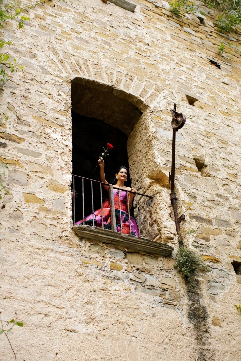 Femminina sorridente in un vestito colorato alzando una rosa rossa, seduta su un balcone di una vecchia casa di pietra con muri beige, in un ambiente rustico con piante verdi vicino alla finestra.