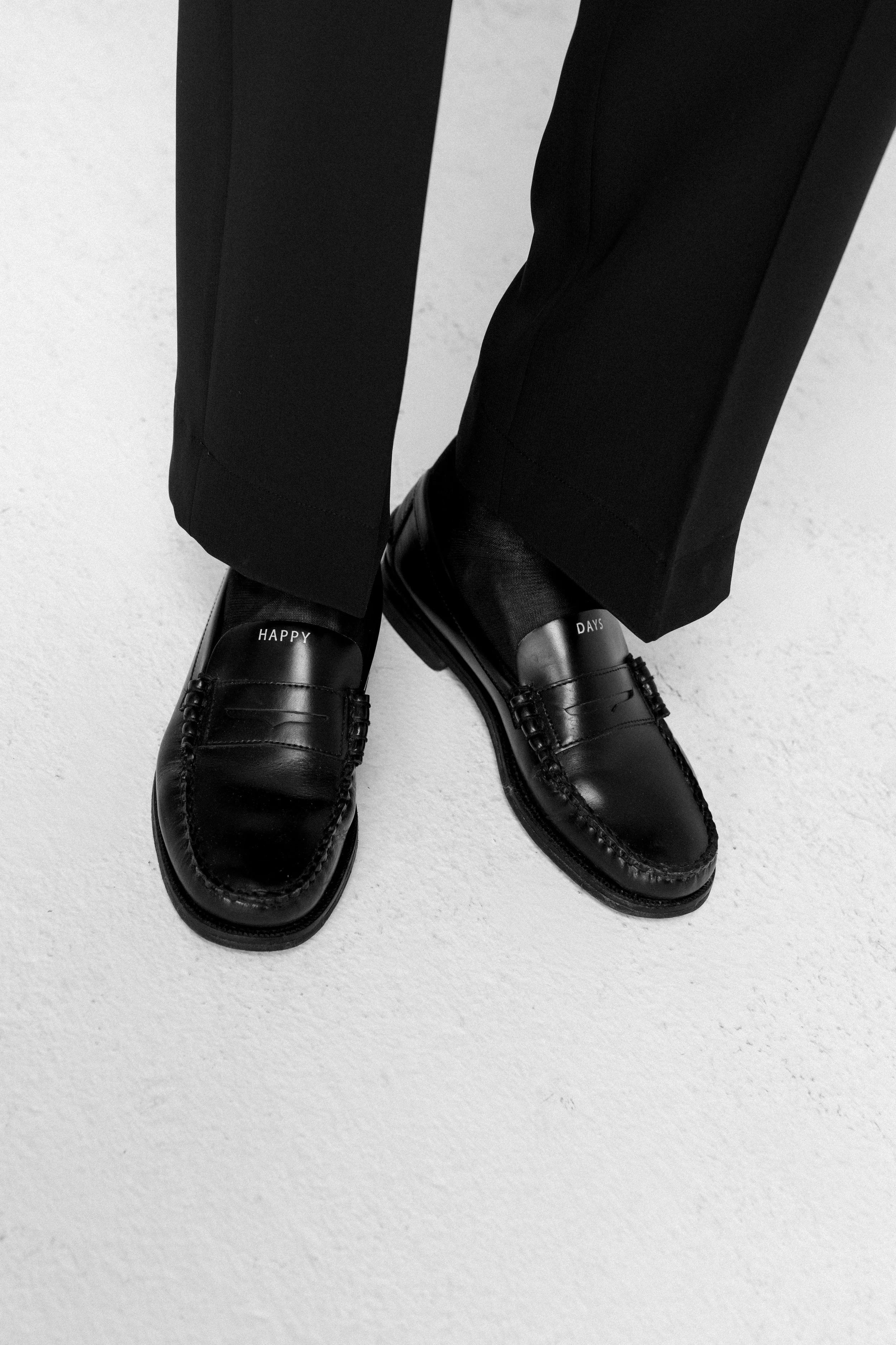 Scarpe nere con scritta bianca 'HAPPY' e 'DAYS' su i piedi di una persona vestita con pantaloni neri, viste dall'alto in bianco e nero.