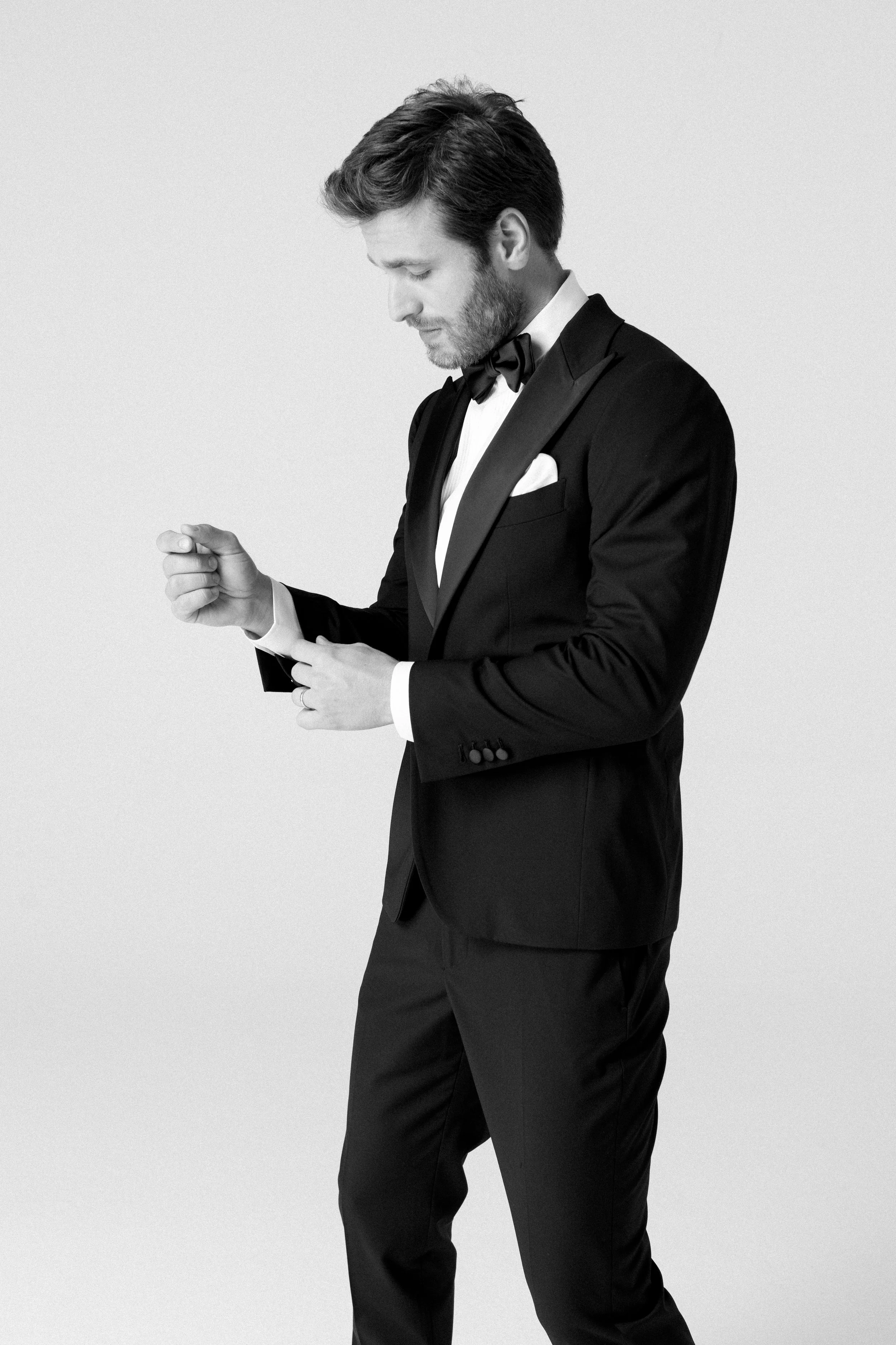 Uomo in abito da sera nero con papillon, regolando i polsini del camice, sfondo bianco