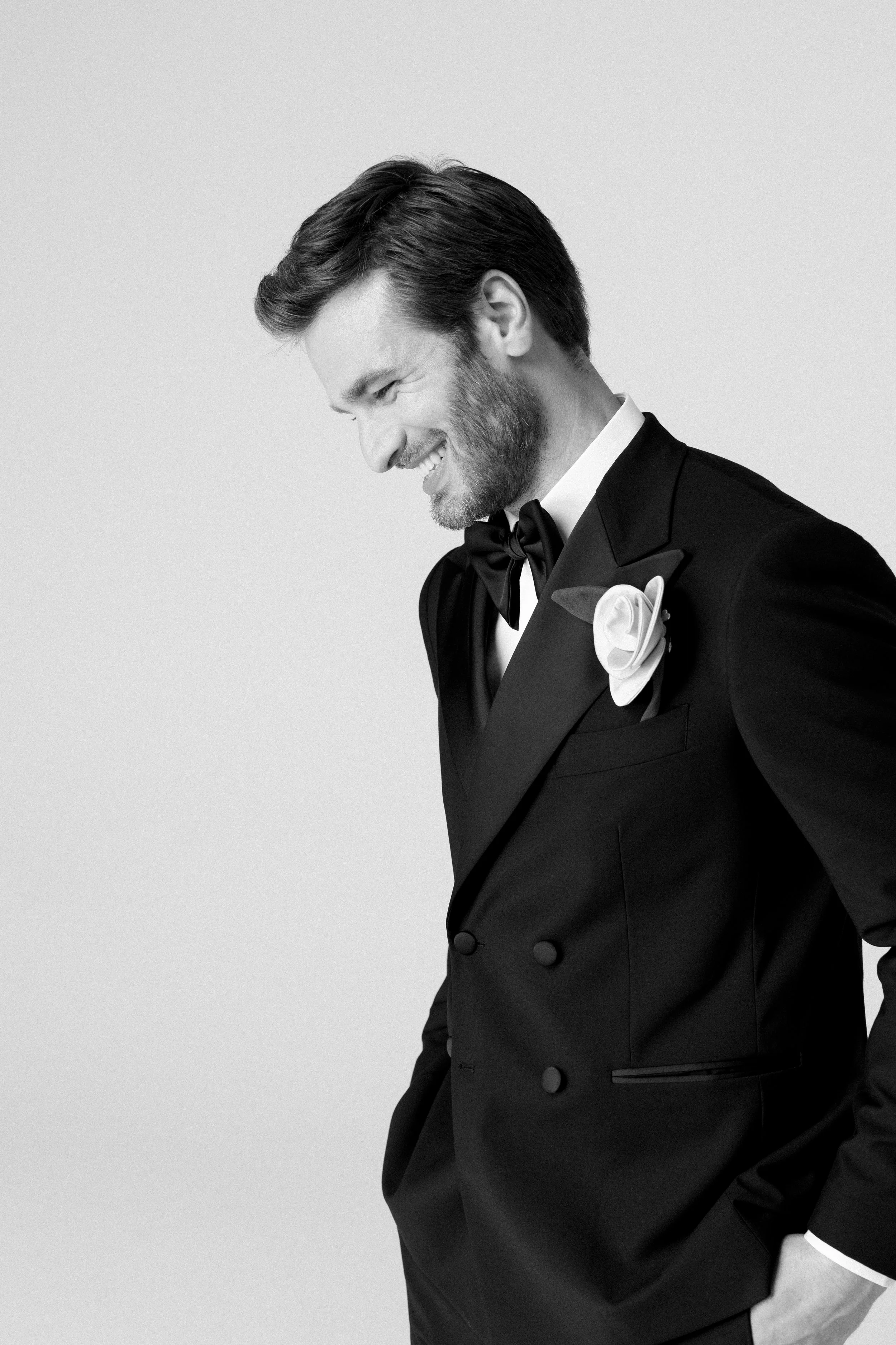 Uomo sorridente indossa un completo da sera nero con papillon e fiore sulla giacca, in bianco e nero.