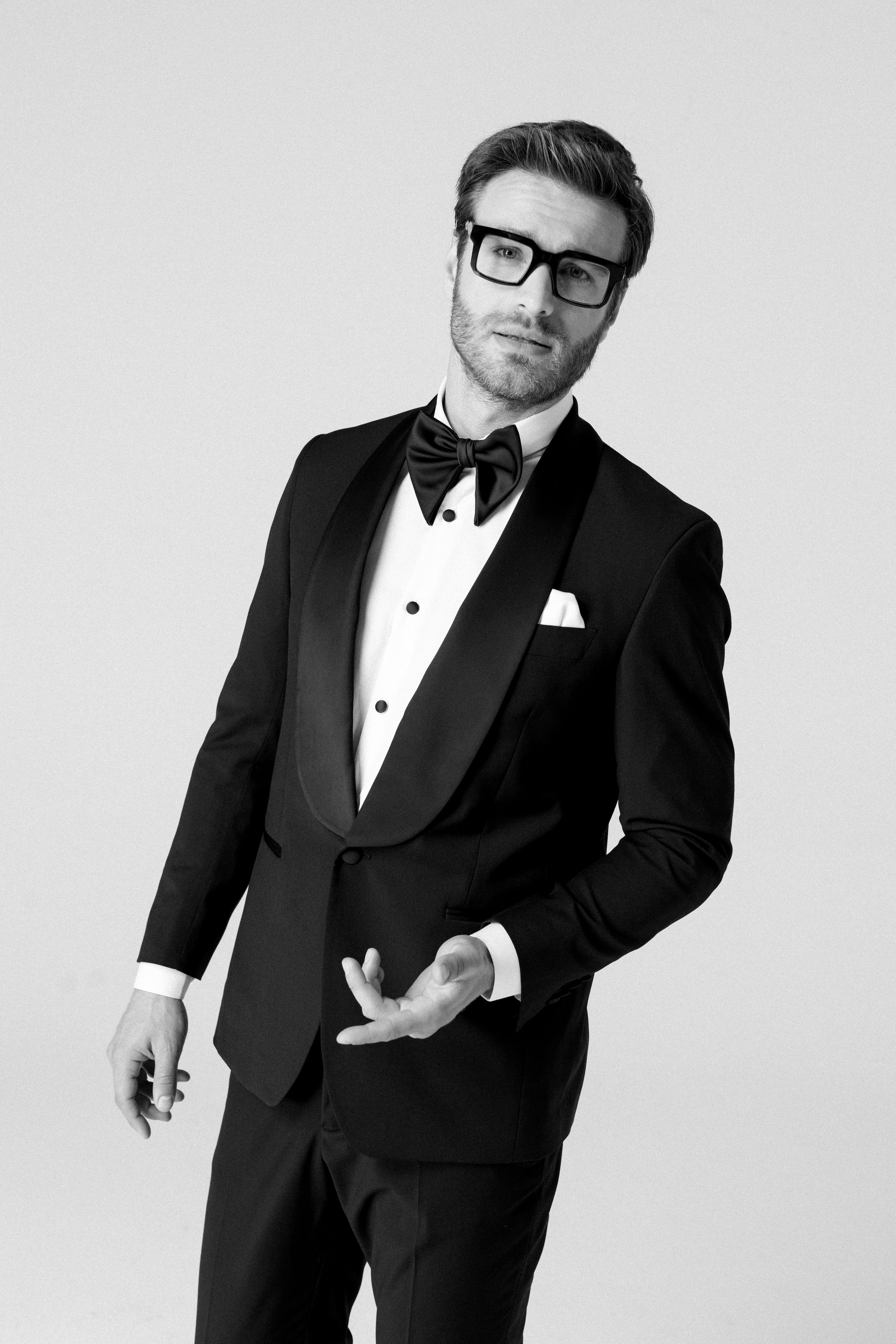 Uomo in abito da sera con occhiali, cravatta a farfalla e giacca nera, posa con espressione curiosa, in bianco e nero.