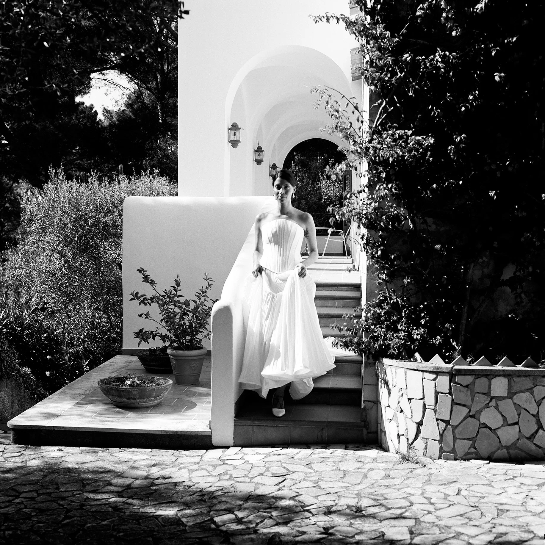 Donna in abito da sposa sulla scalinata di un edificio bianco con archi, circondata da piante.