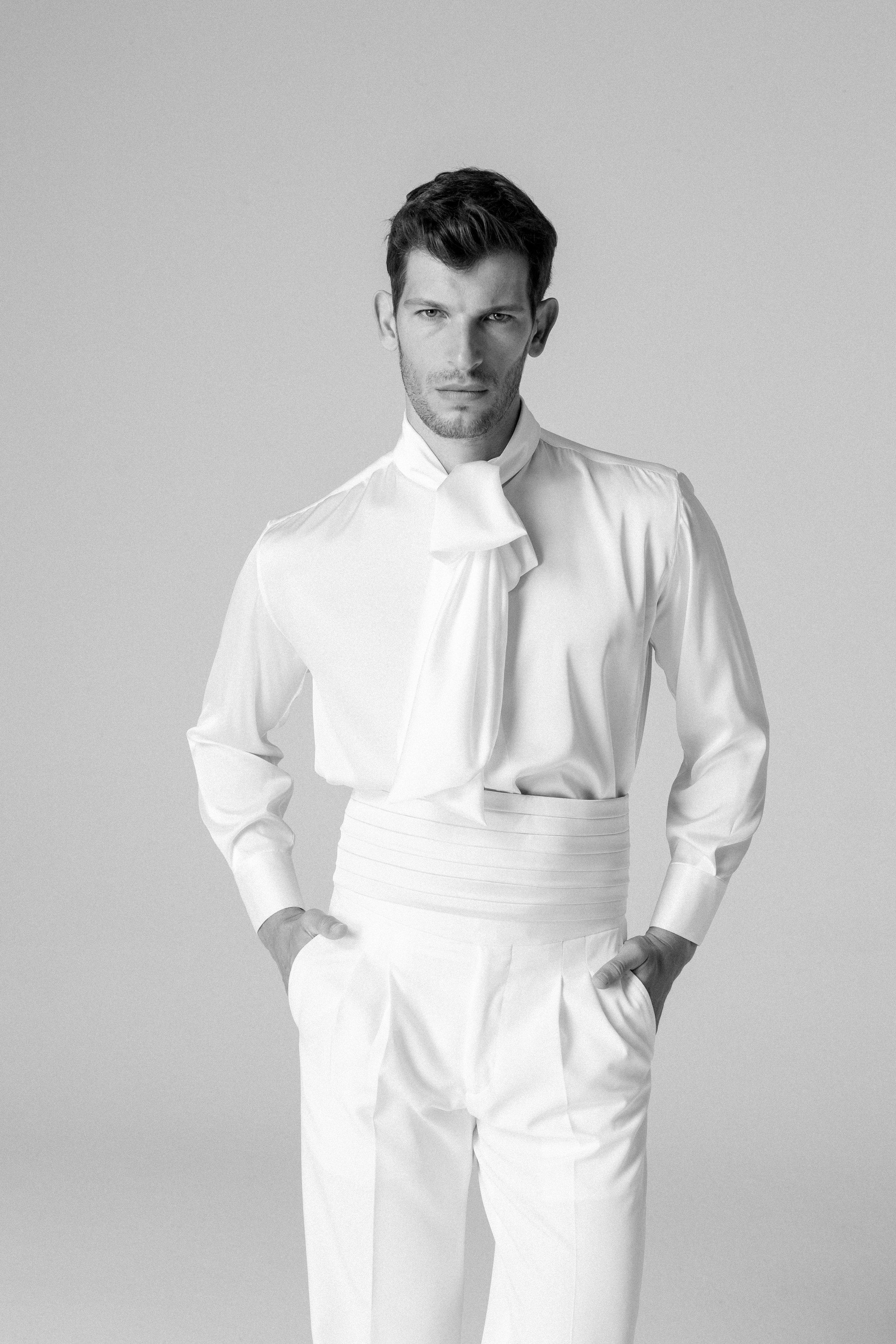 Uomo elegante in abbigliamento bianco con cravatta da papillon, posa con le mani in tasca, sfondo semplice.