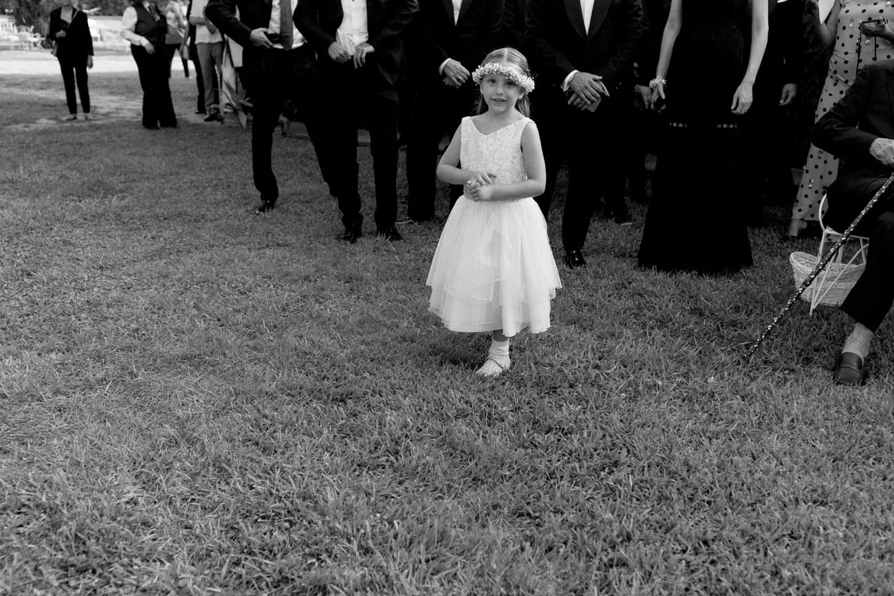 Una giovane ragazza con un vestito bianco e una corona di fiori sulla testa, che cammina su un prato durante un evento, mentre sullo sfondo ci sono persone vestite elegantemente.