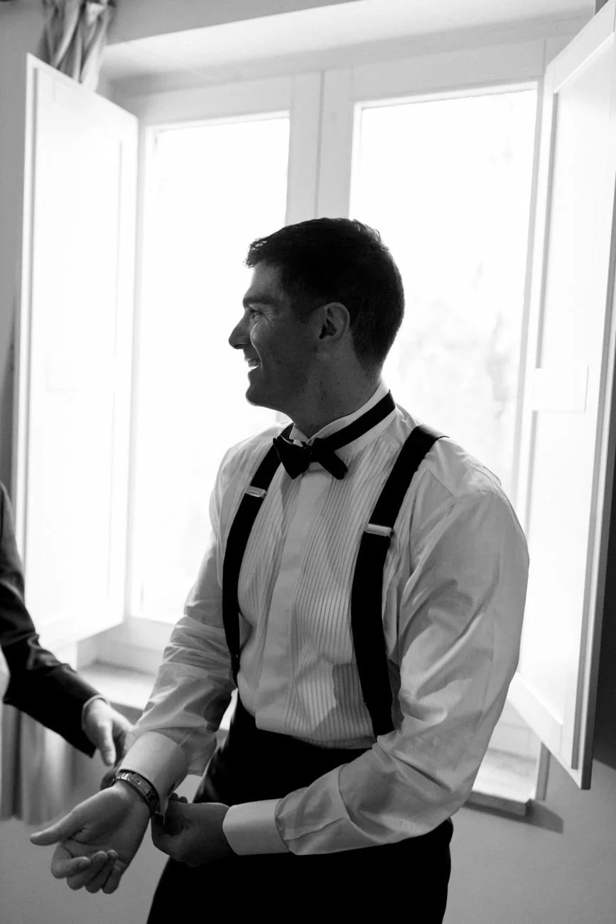 Uomo sorridente con abito elegante, camicia bianca con plissettature, papillon nero e bretelle, in un ambiente luminoso originale con finestre aperte.