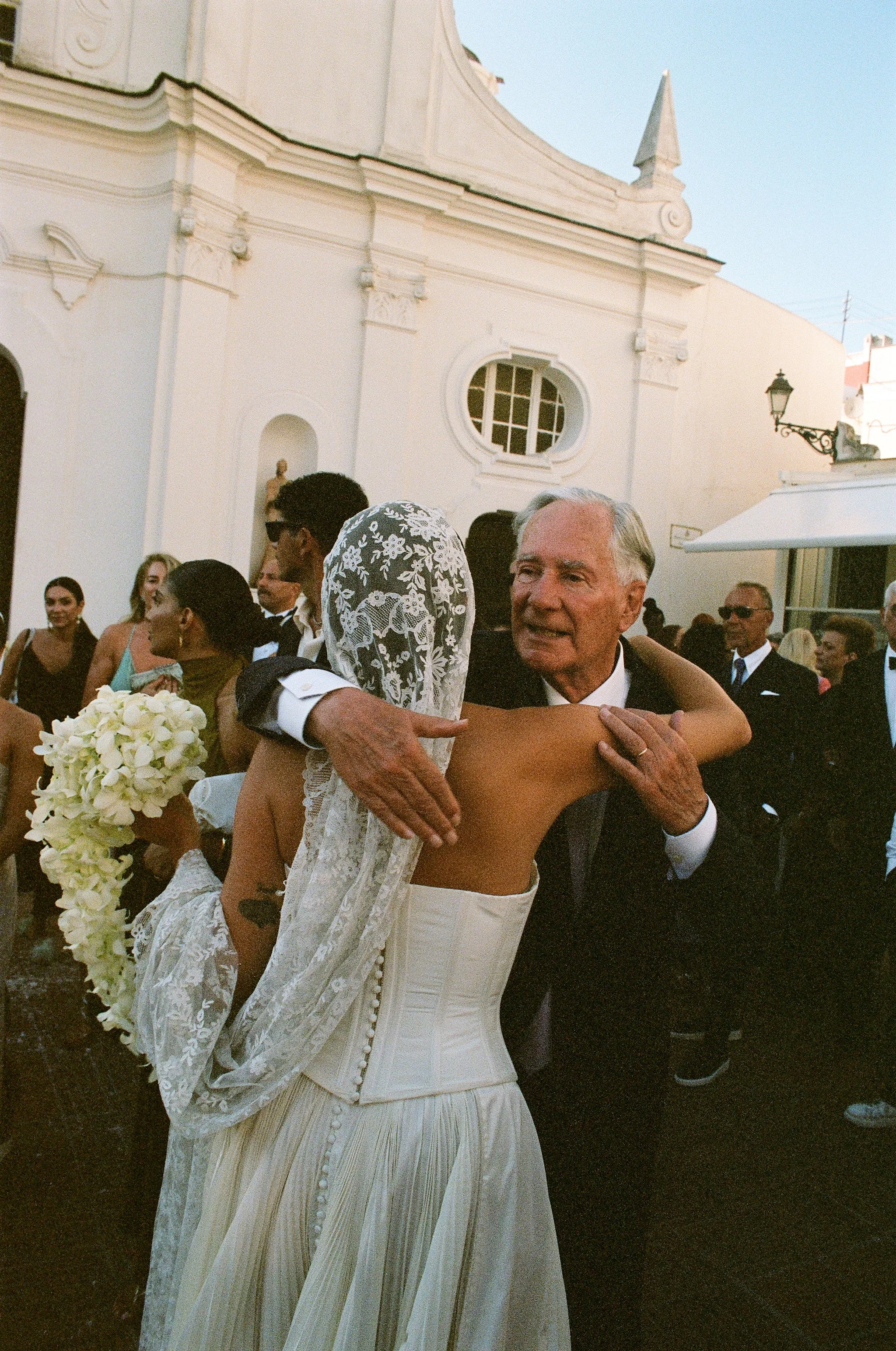 Spose in un matrimonio con una donna vestita in abito bianco e un uomo anziano che la abbraccia, anche altre persone vestite elegantemente si trovano sullo sfondo all'aperto.