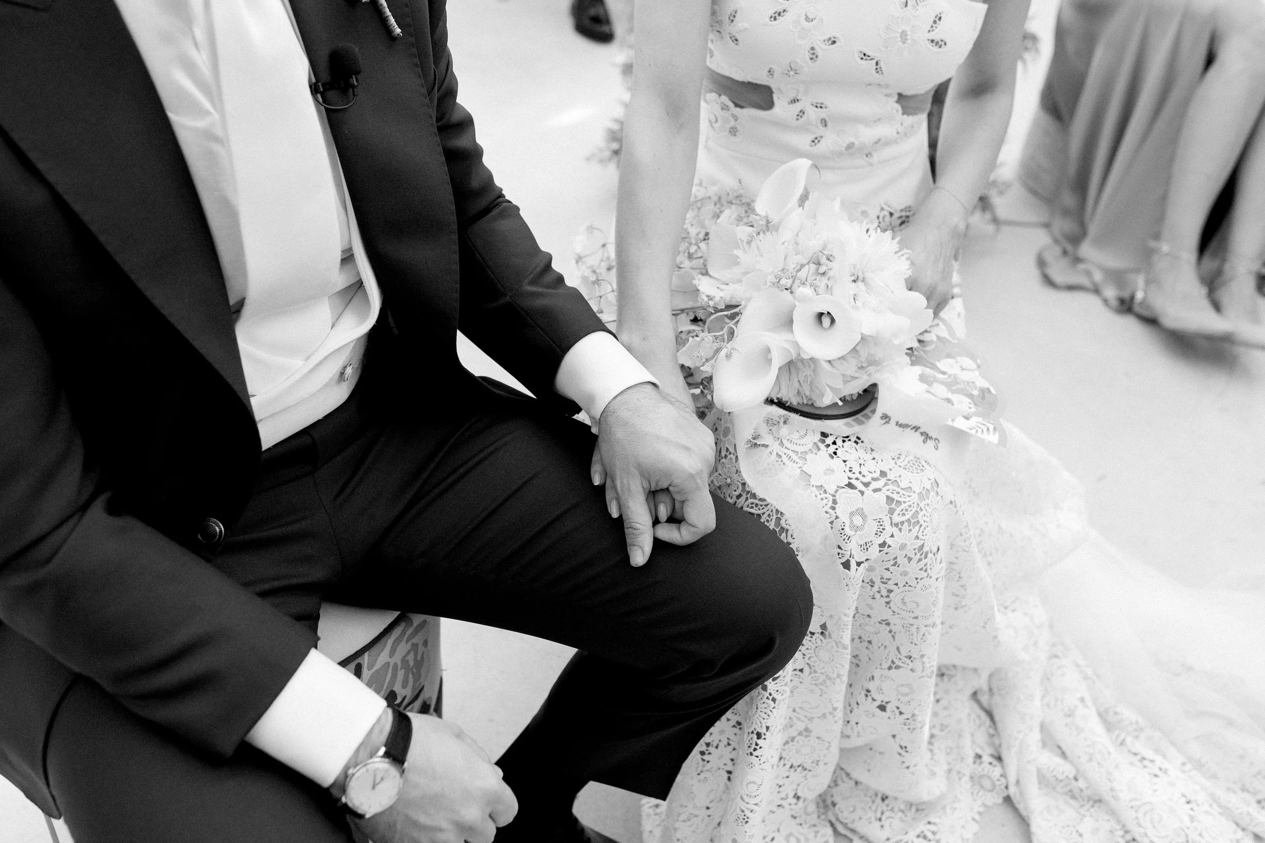 Coppia di sposi seduti, si tengono per mano, il primo indossa un abito nero e una camicia bianca, la sposa indossa un abito da sposa con ricami e tiene un bouquet di fiori bianchi.