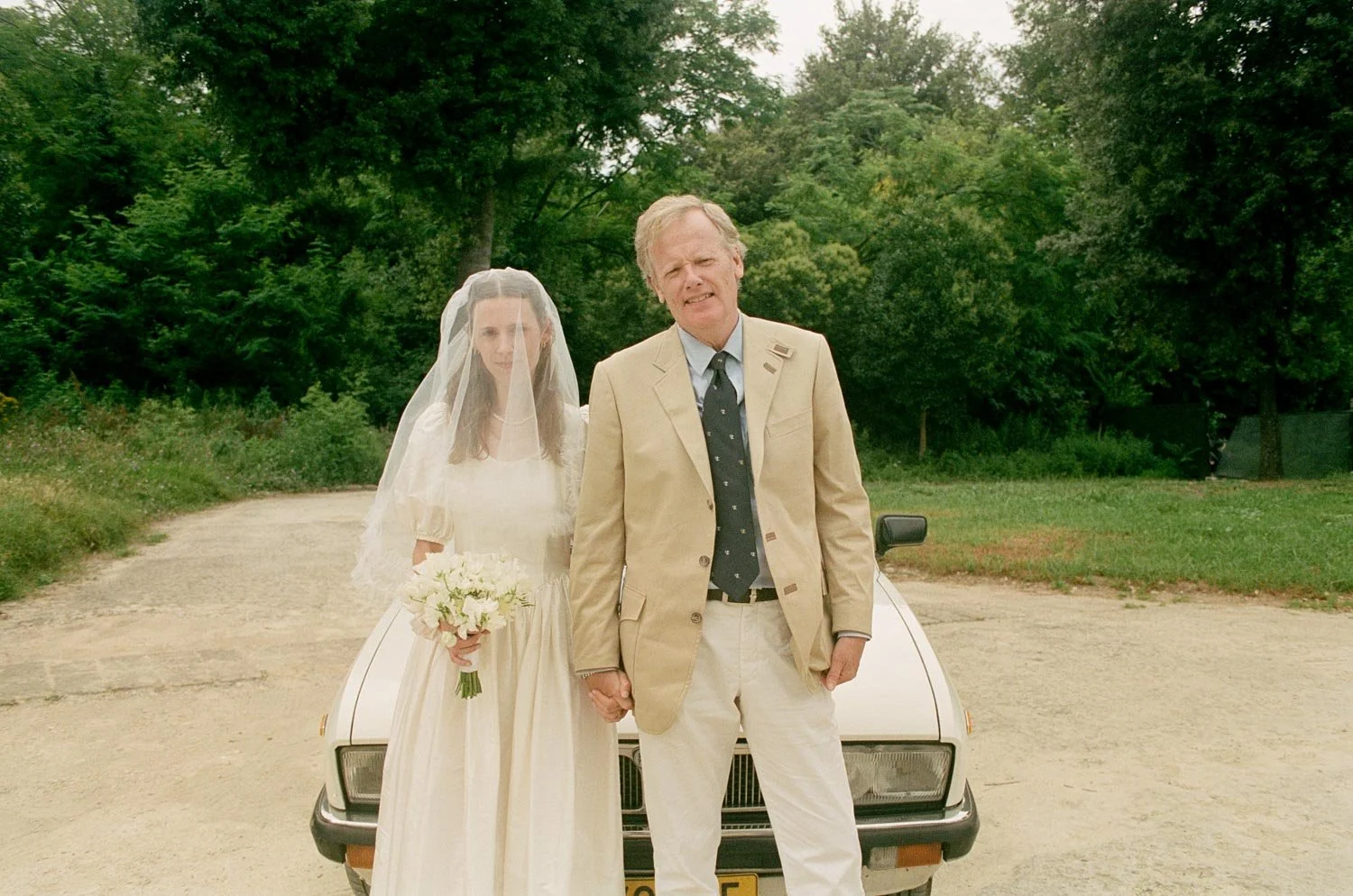 Un uomo e una donna in abiti da matrimonio si tengono per mano davanti a un'auto, con uno sfondo di alberi e natura.
