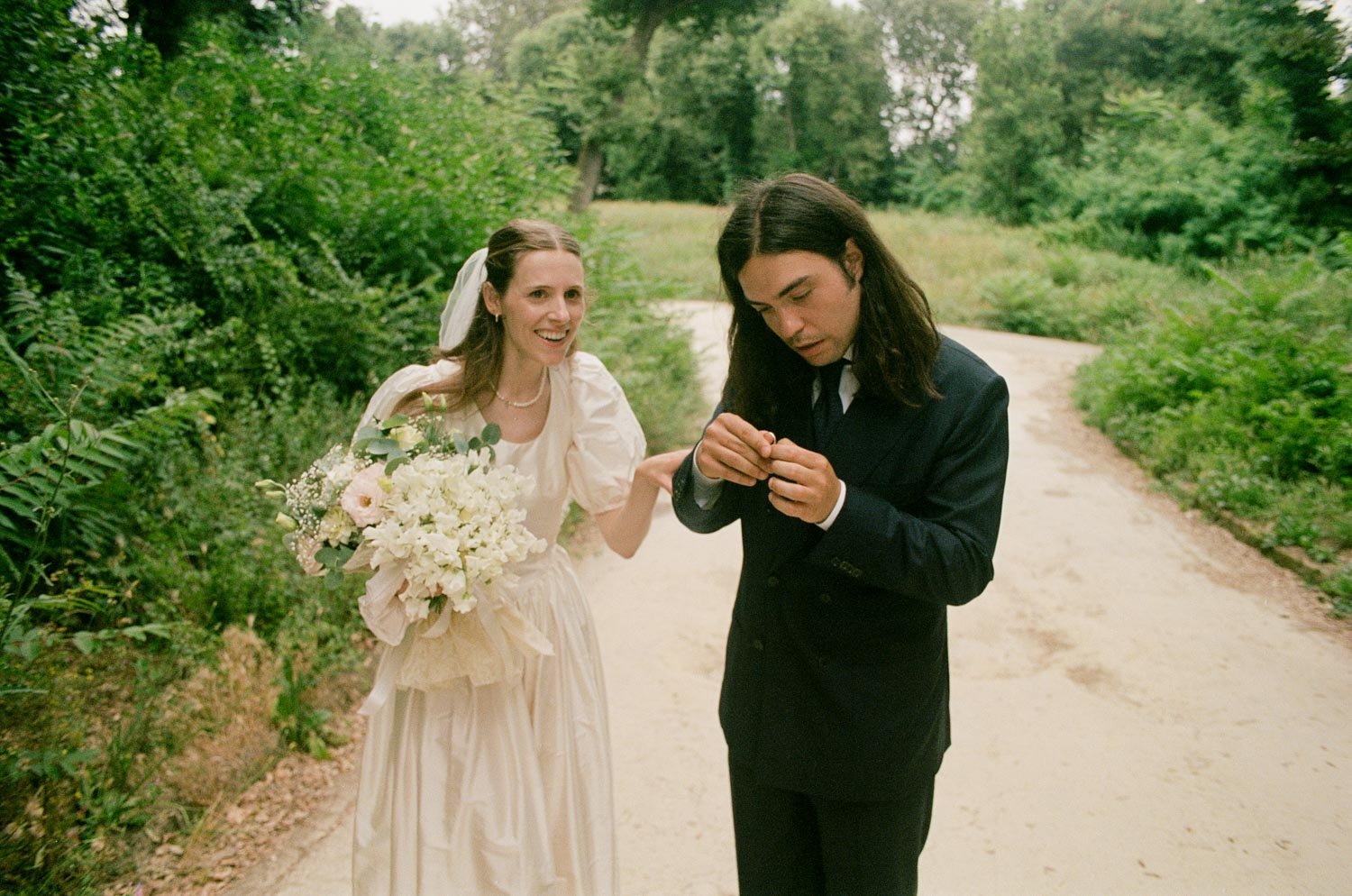 Una coppia di persone, una donna vestita da sposa e un uomo in abito, si trovano su un sentiero nel verde. La donna sorride mentre l'uomo guarda un oggetto in mano.