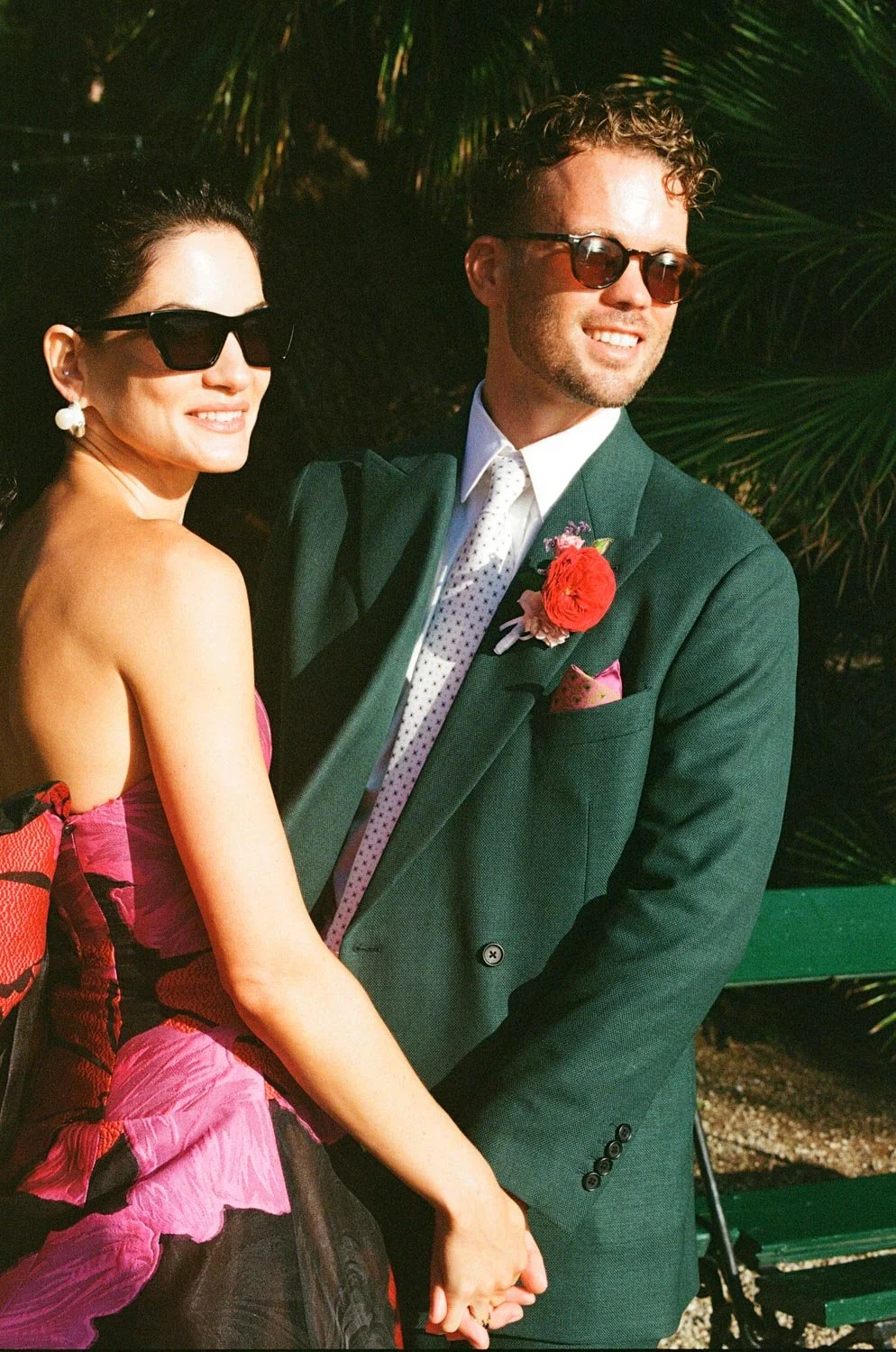 Una coppia elegante indossa occhiali da sole e si tiene per mano, sorridendo in un ambiente all'aperto con pianta di palme sullo sfondo.