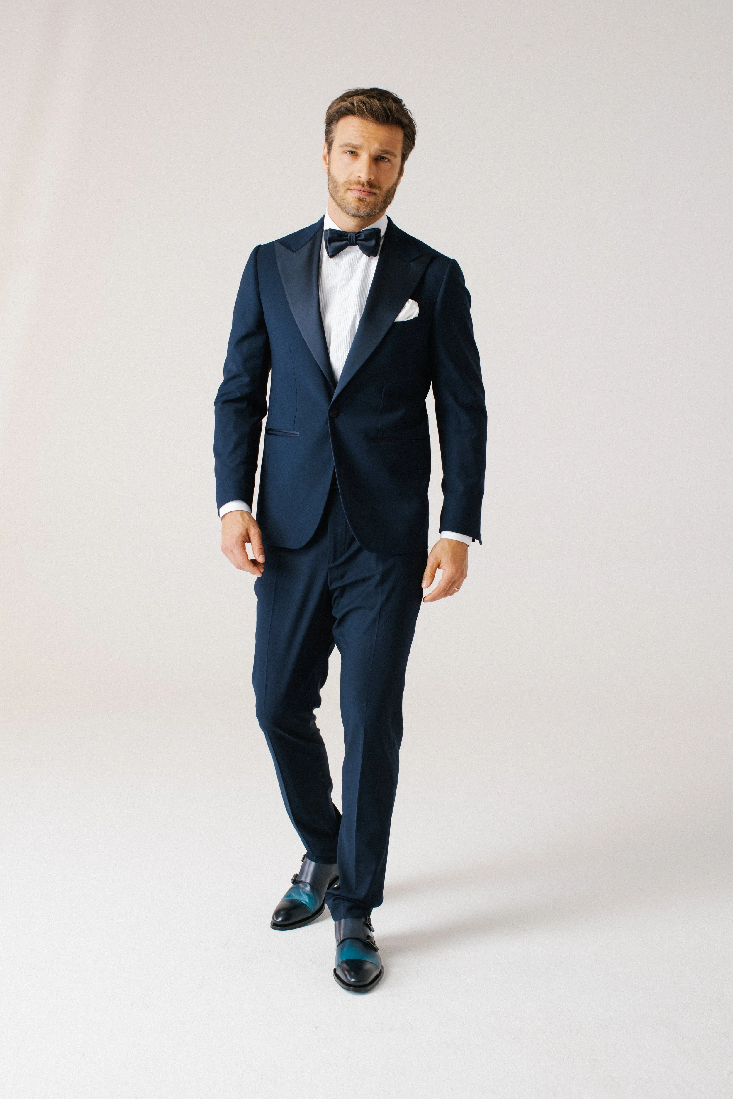 Uomo elegante vestito con completo blu, fiocco e scarpe nere, che cammina.