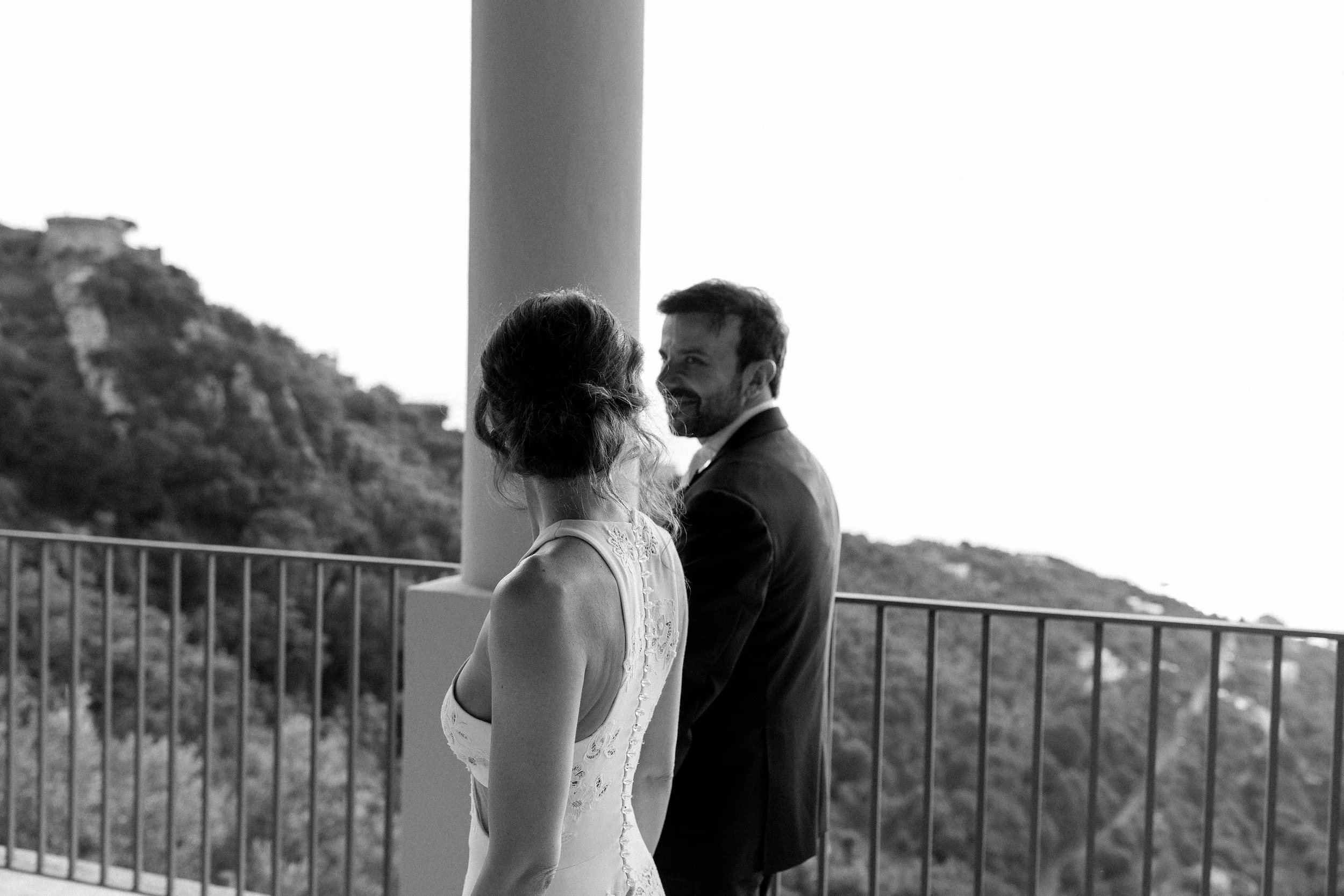 Una coppia durante un matrimonio, vista di profilo di una donna con abito da sposa e un uomo in abito da cerimonia, su un balcone con vista su una collina e un mare lontano.