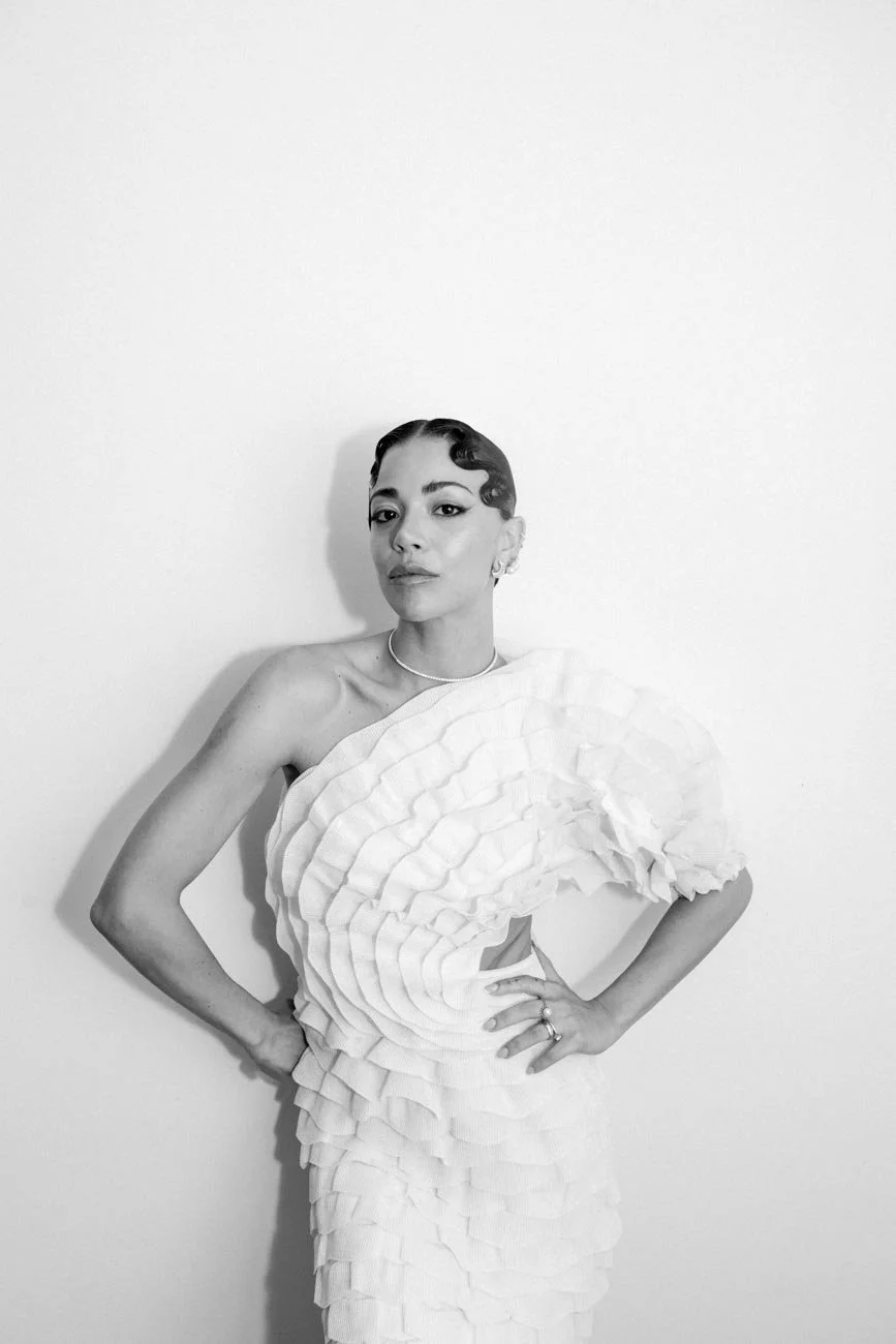 Donna con capelli corti e riccioli, indossa un abito bianco elegante con dettagli a onde, posa contro una parete bianca.