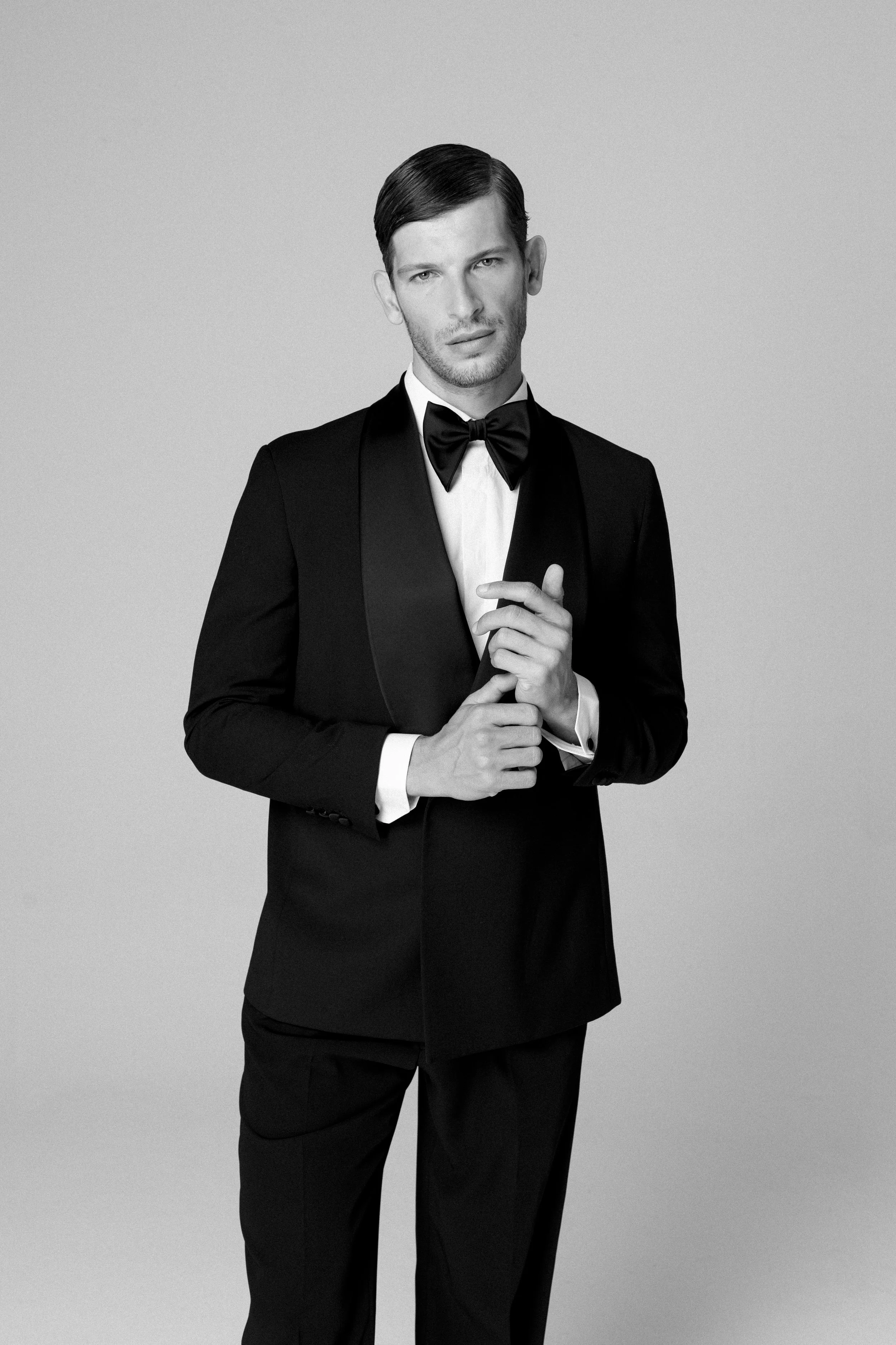 Uomo in abito da sera nero con papillon, posa con le mani sfiorandosi, sfondo semplice.
