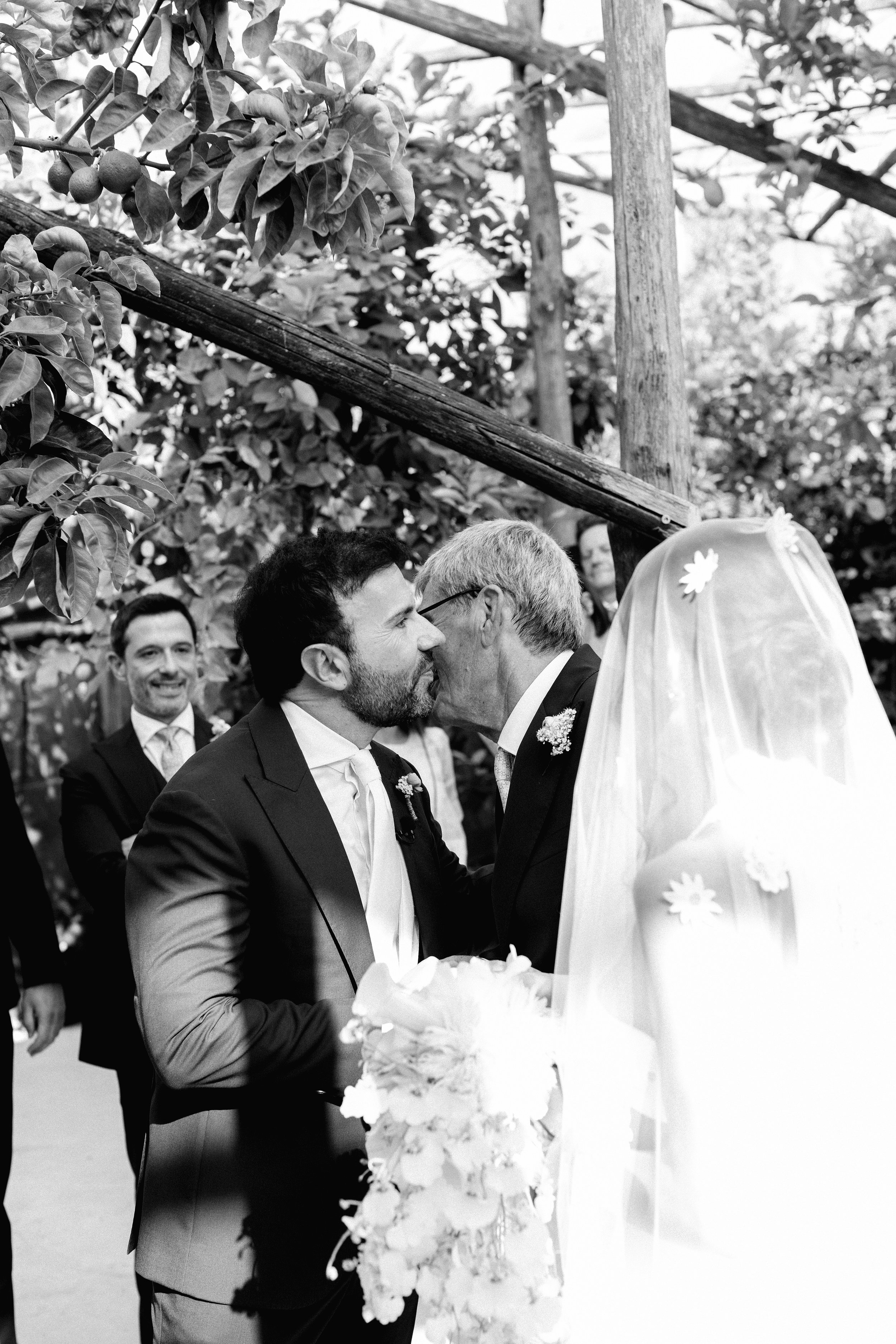 Un matrimonio tra due uomini si bacia durante una cerimonia all'aperto, con altri uomini sorridenti sullo sfondo.