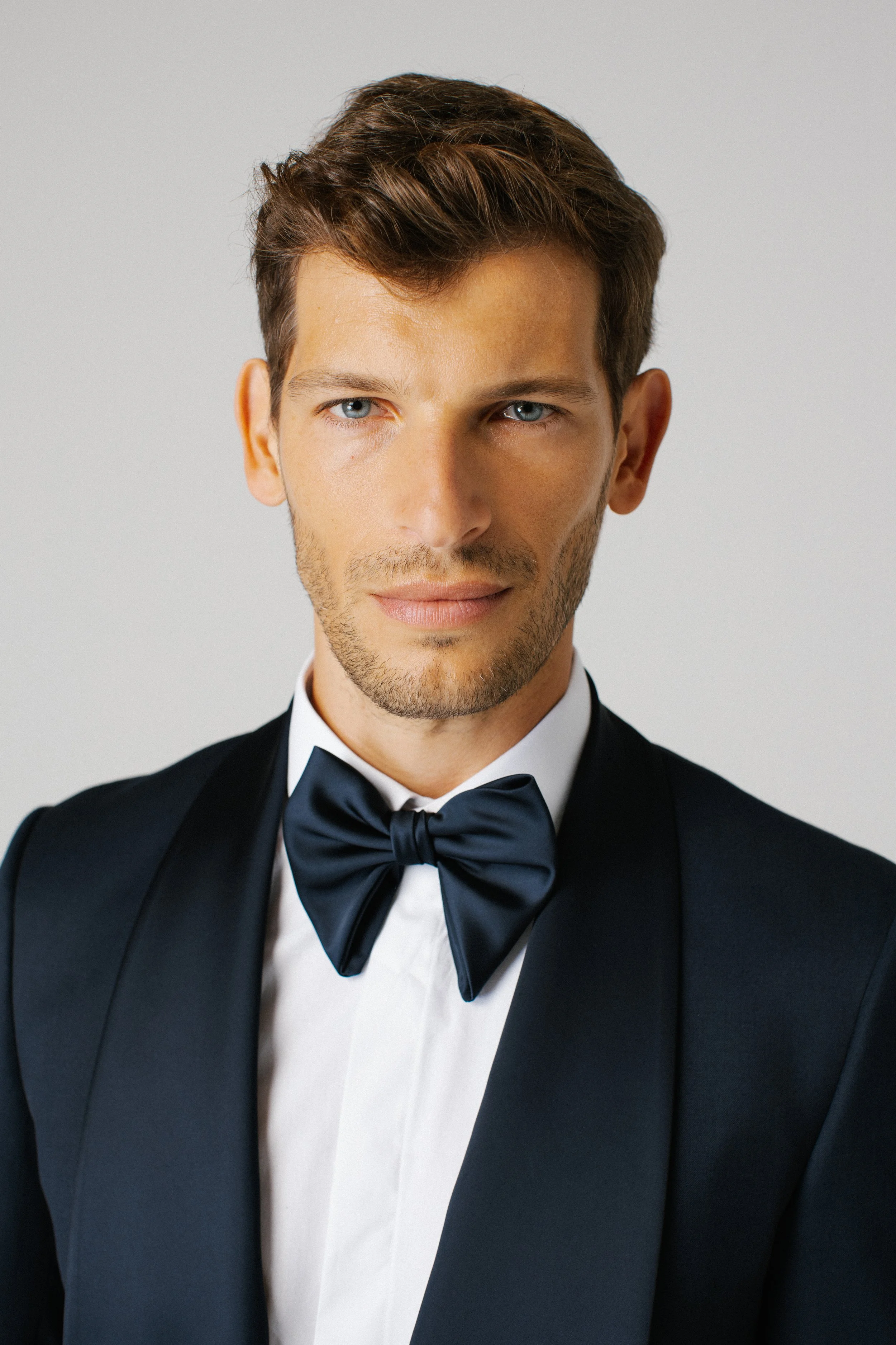 Uomo elegante con abito da sera nero e papillon, sorriso sottile, sfondo grigio chiaro.