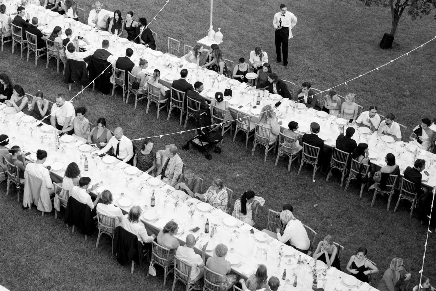 Un'immagine all'aperto di lunga tavolata con persone sedute che partecipano a un evento sociale, sotto una tenda con luci decorative e un operatore che tiene una bottiglia di vino.