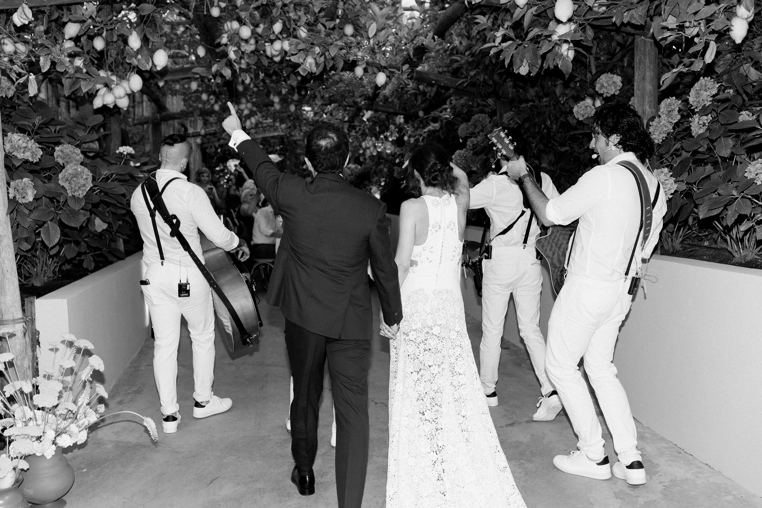 Una coppia di sposi, una donna in abito bianco lungo e un uomo in completo nero, camminano mano nella mano attraverso un'area decorata con piante e fiori, accompagnati da musicisti e un fotografo.