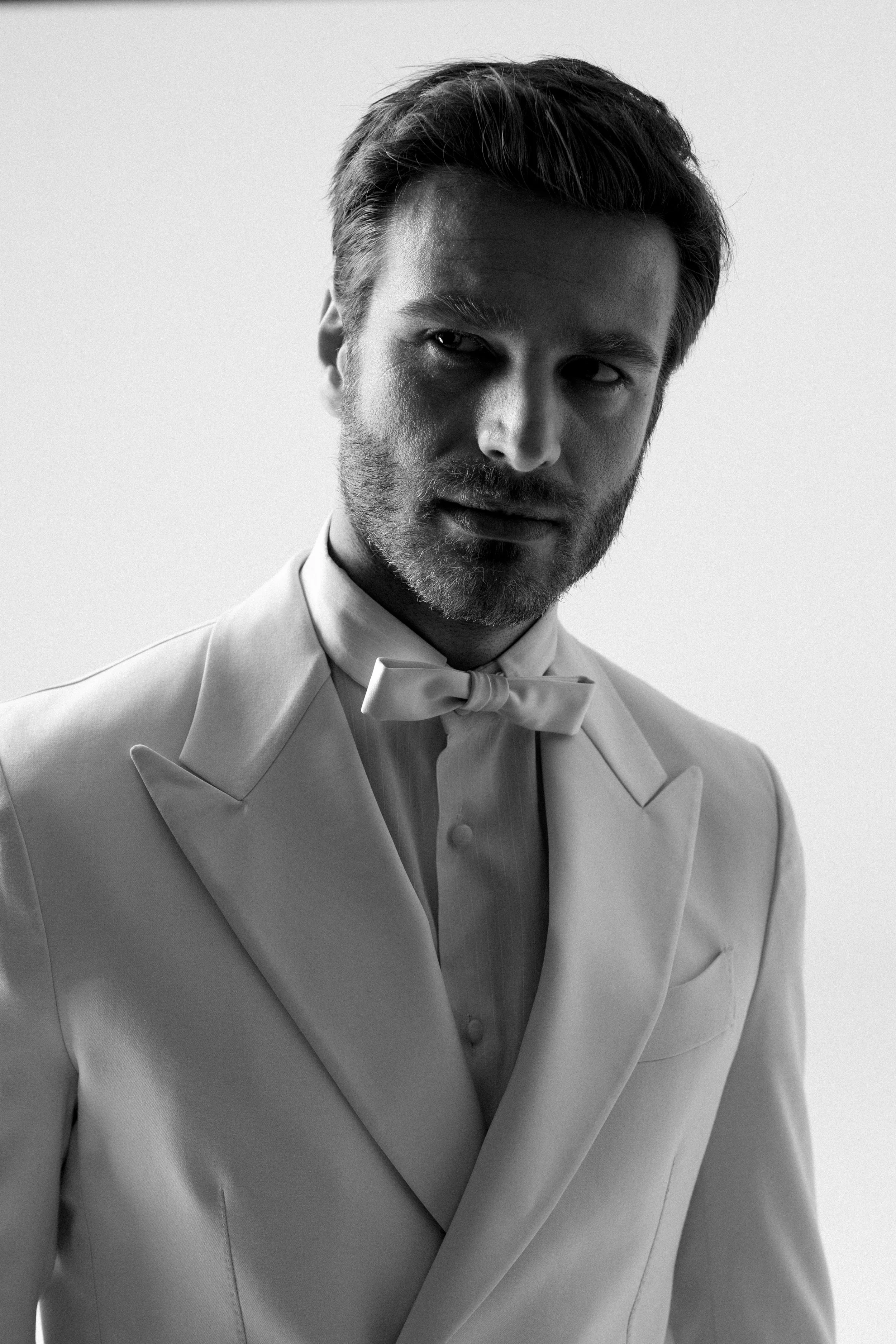 Ritratto di un uomo con baffi e barba, vestito con un completo bianco e un papillon, in bianco e nero.