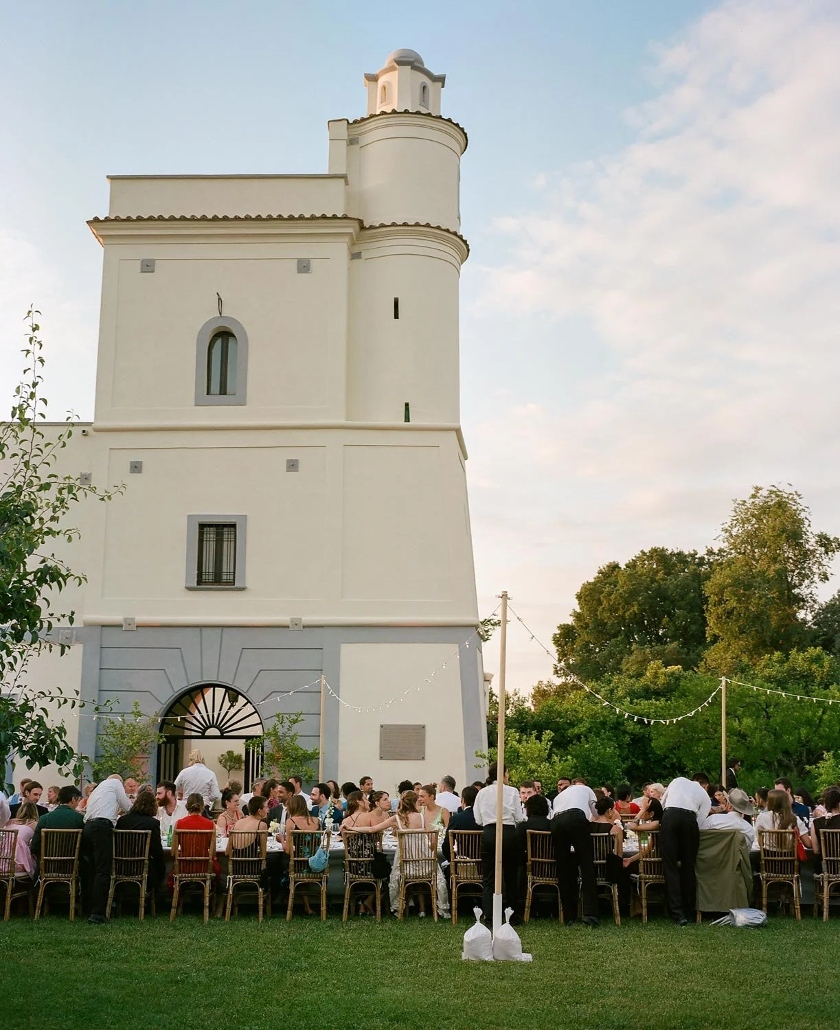 Un gruppo di persone sedute a tavola all'aperto davanti a un grande edificio bianco con una torre, probabilmente durante un evento sociale o una celebrazione serale.