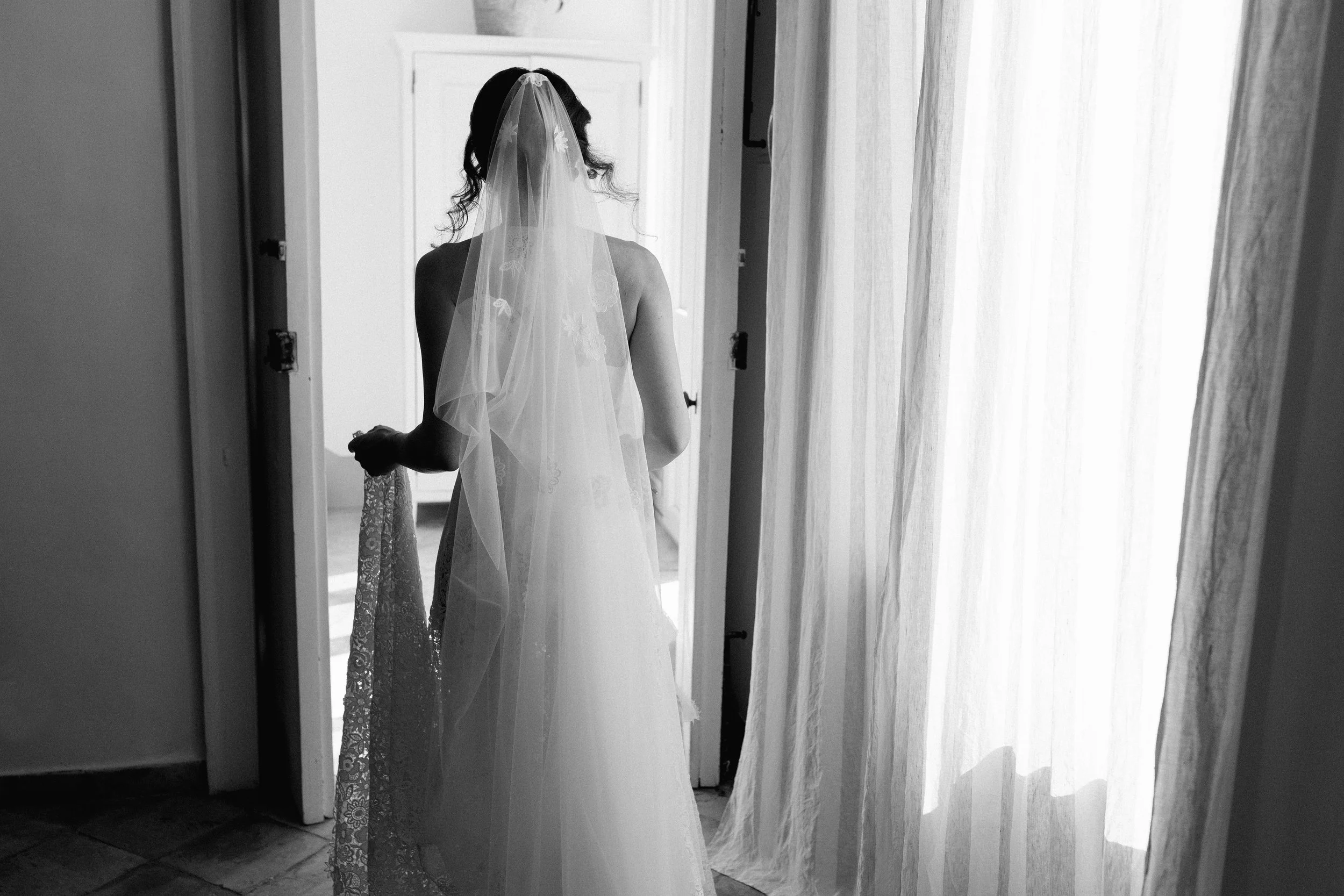 Una donna con un abito da sposa e un velo bianco si trova in una stanza, vista da dietro, che si affaccia verso una porta aperta. La stanza è illuminata dalla luce naturale attraverso le tende trasparenti.