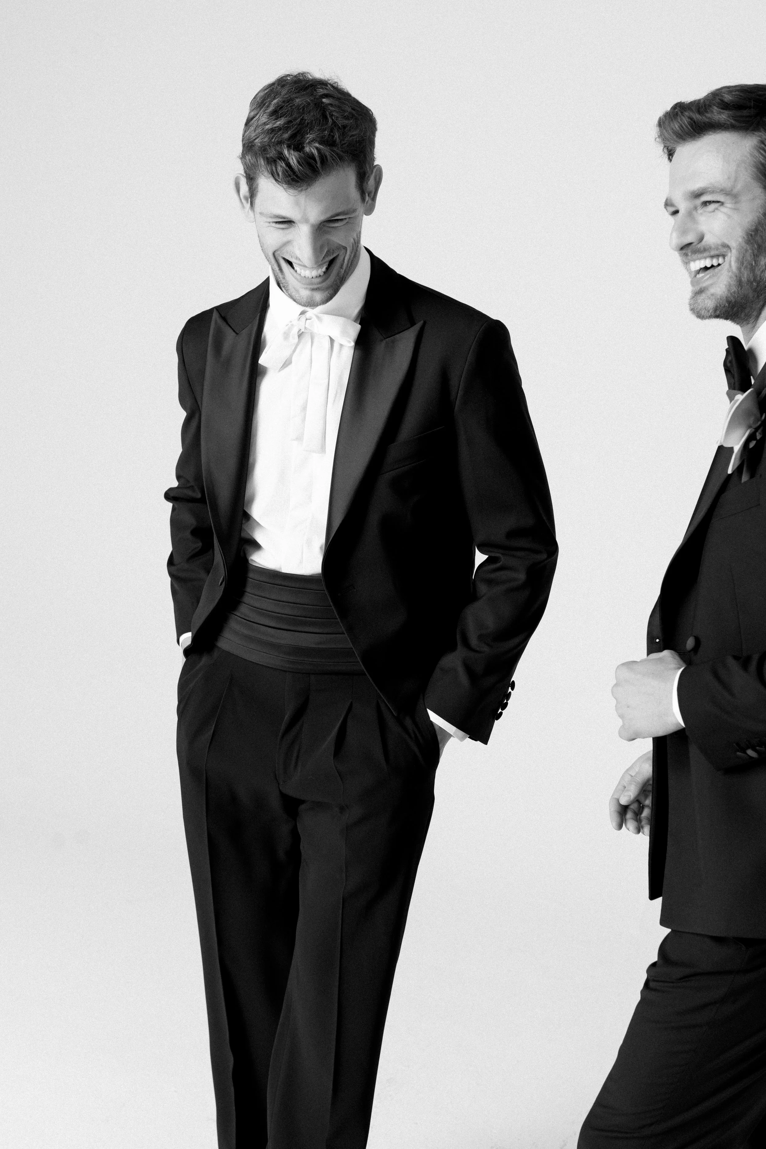 Due uomini sorridono e ridono mentre indossano abiti formali nero e bianco, in una foto in bianco e nero contro uno sfondo chiaro.