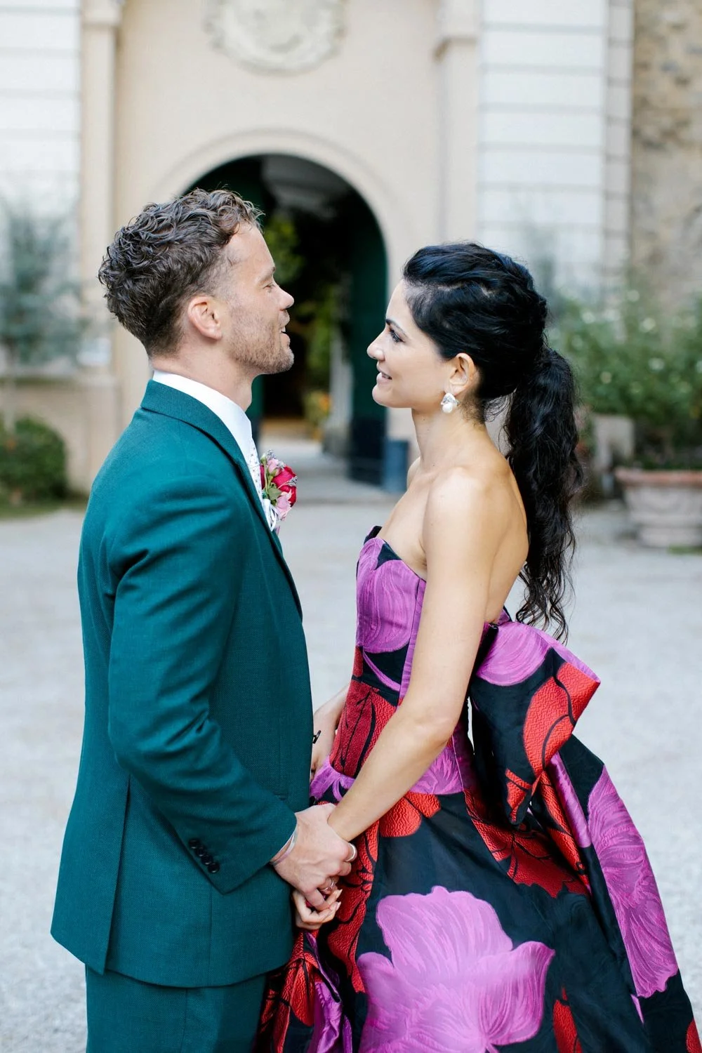 Una coppia infila le mani tra loro, sorridendosi, all'esterno davanti a un edificio storico di pietra e mattoni, con vegetazione sullo sfondo.