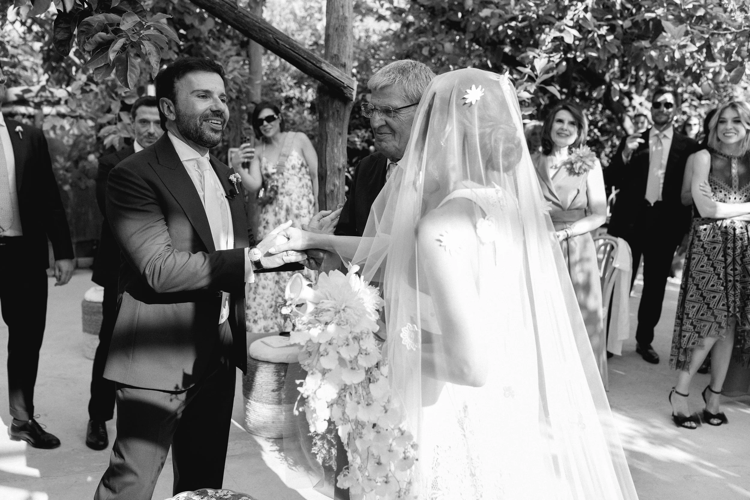 Scena di un matrimonio, con il fidanzato che tiene le mani della sposa durante la cerimonia all'aperto, circondati da amici e parenti sorridenti.