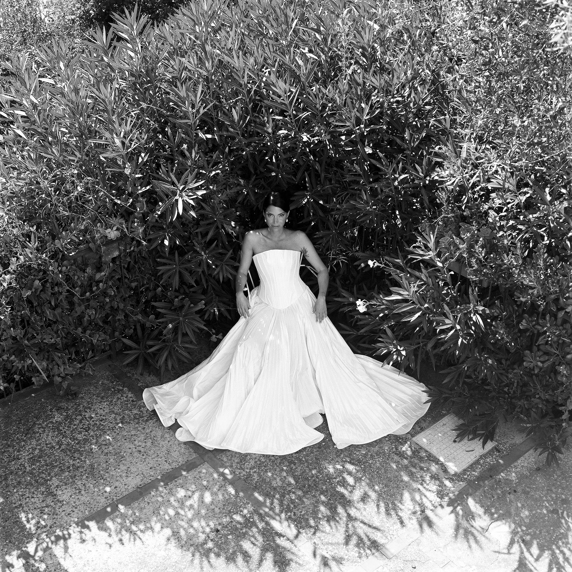 Una donna in abito da sposa bianco si trova davanti a una parete di foglie e piante, con un pavimento di pietra.