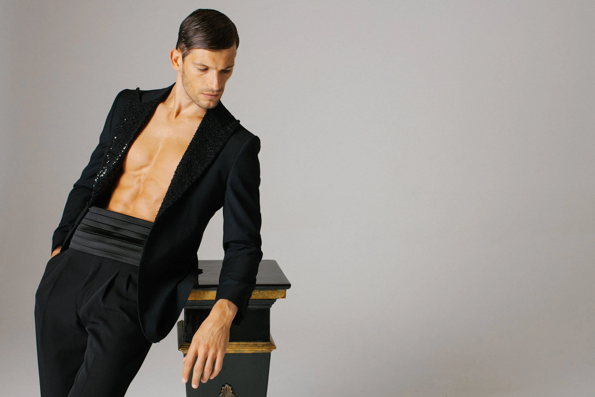 Uomo in abito nero aperto, con addome visibile, appoggiato a un mobile contro uno sfondo grigio.