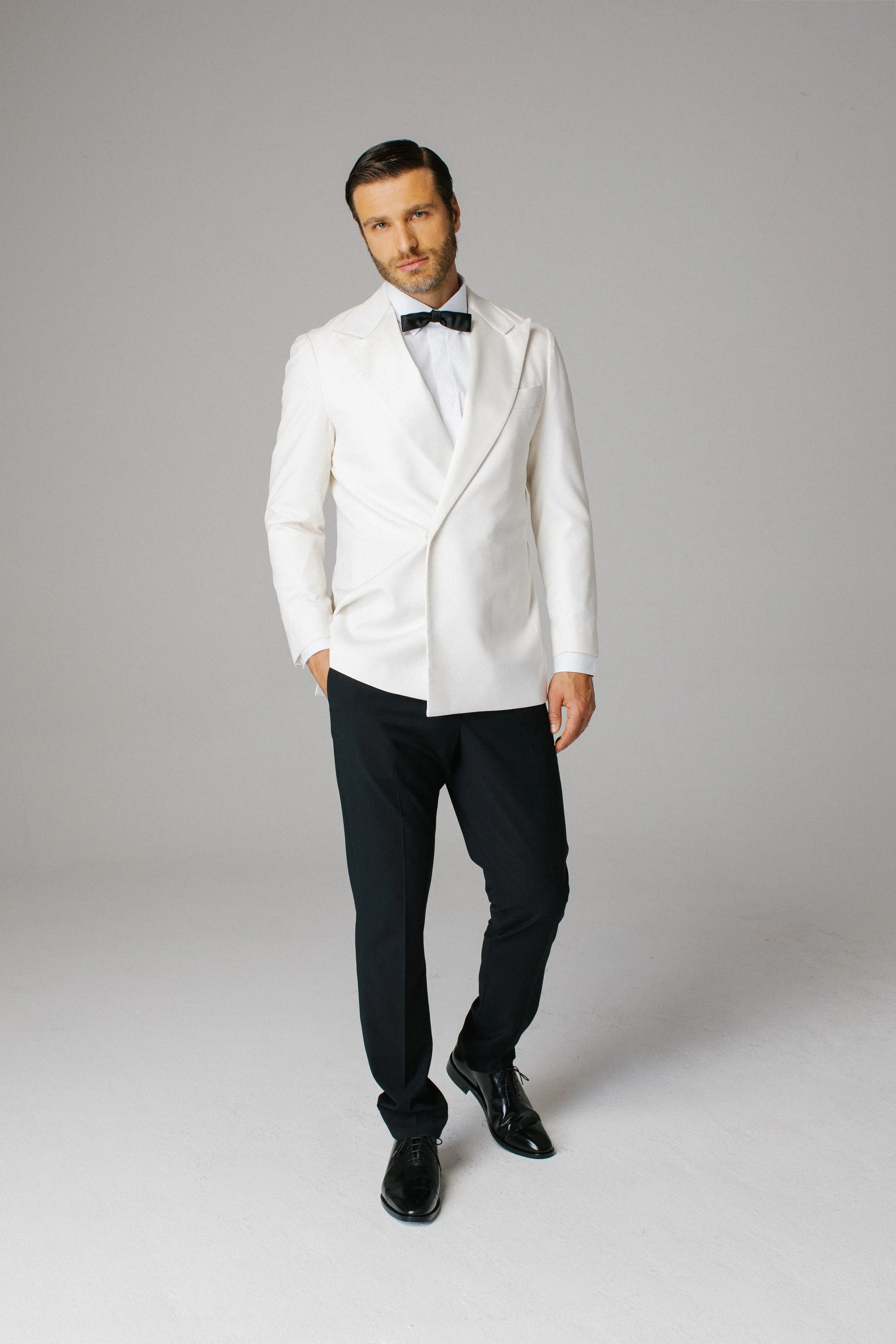 Uomo elegante con abito bianco, papillon nero, pantaloni neri e scarpe lucide, posando contro sfondo grigio.