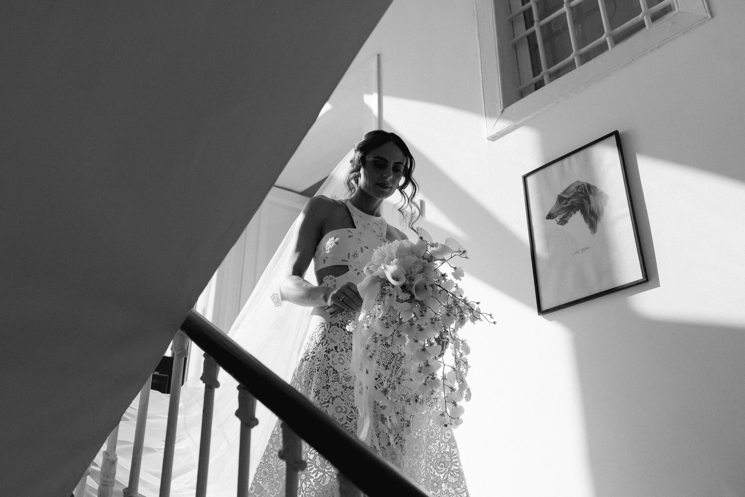 Una sposa in abito di pizzo bianco con un bouquet di fiori, vista di lato dalla scale, in un ambiente con pareti bianche e una finestra con grate, in bianco e nero.