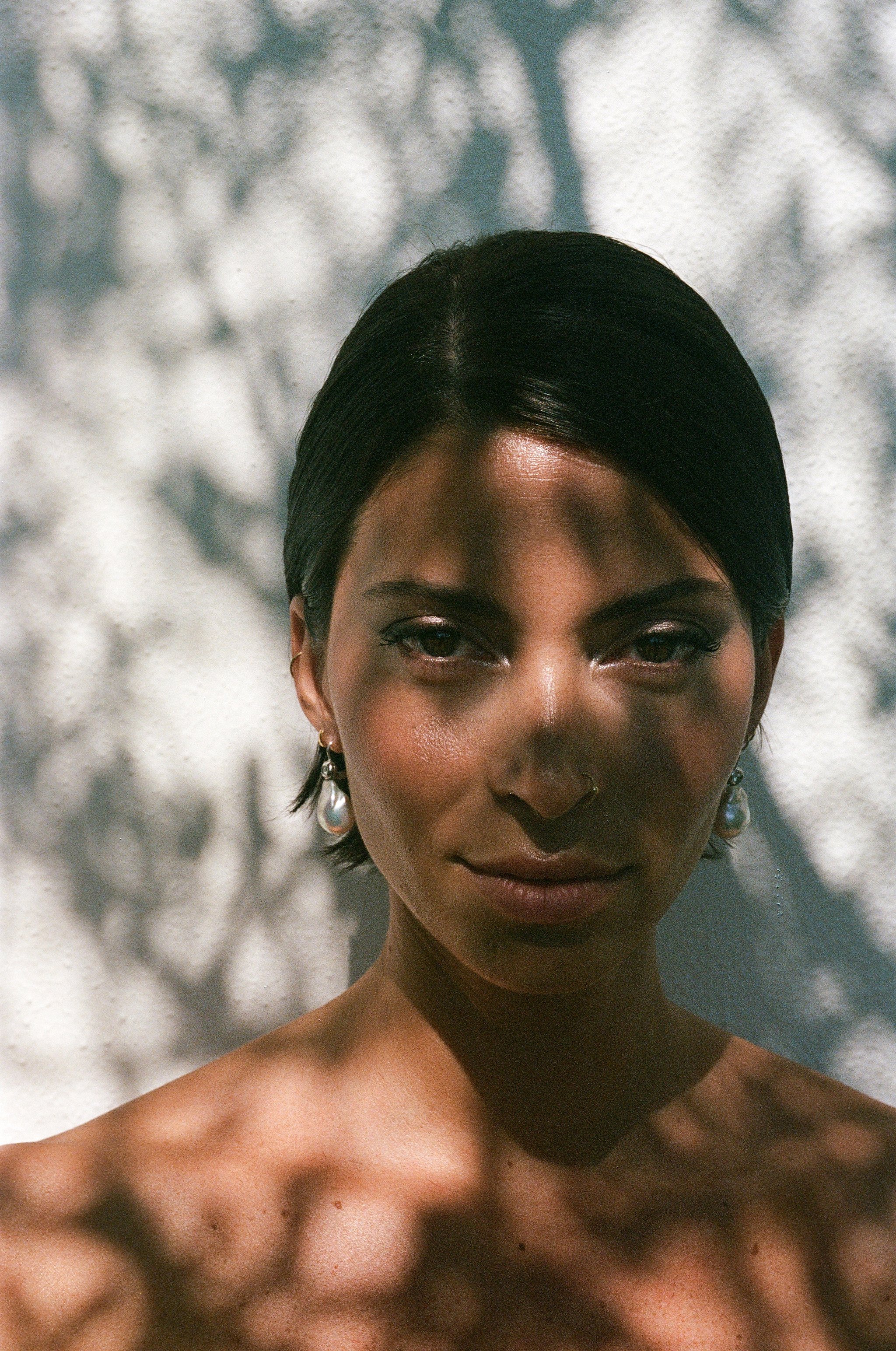 Ritratto di una donna con capelli neri pettinati a scatola, aspettiatrice di luci e ombre proiettate su volto e spalle, con orecchini di perle pendenti.