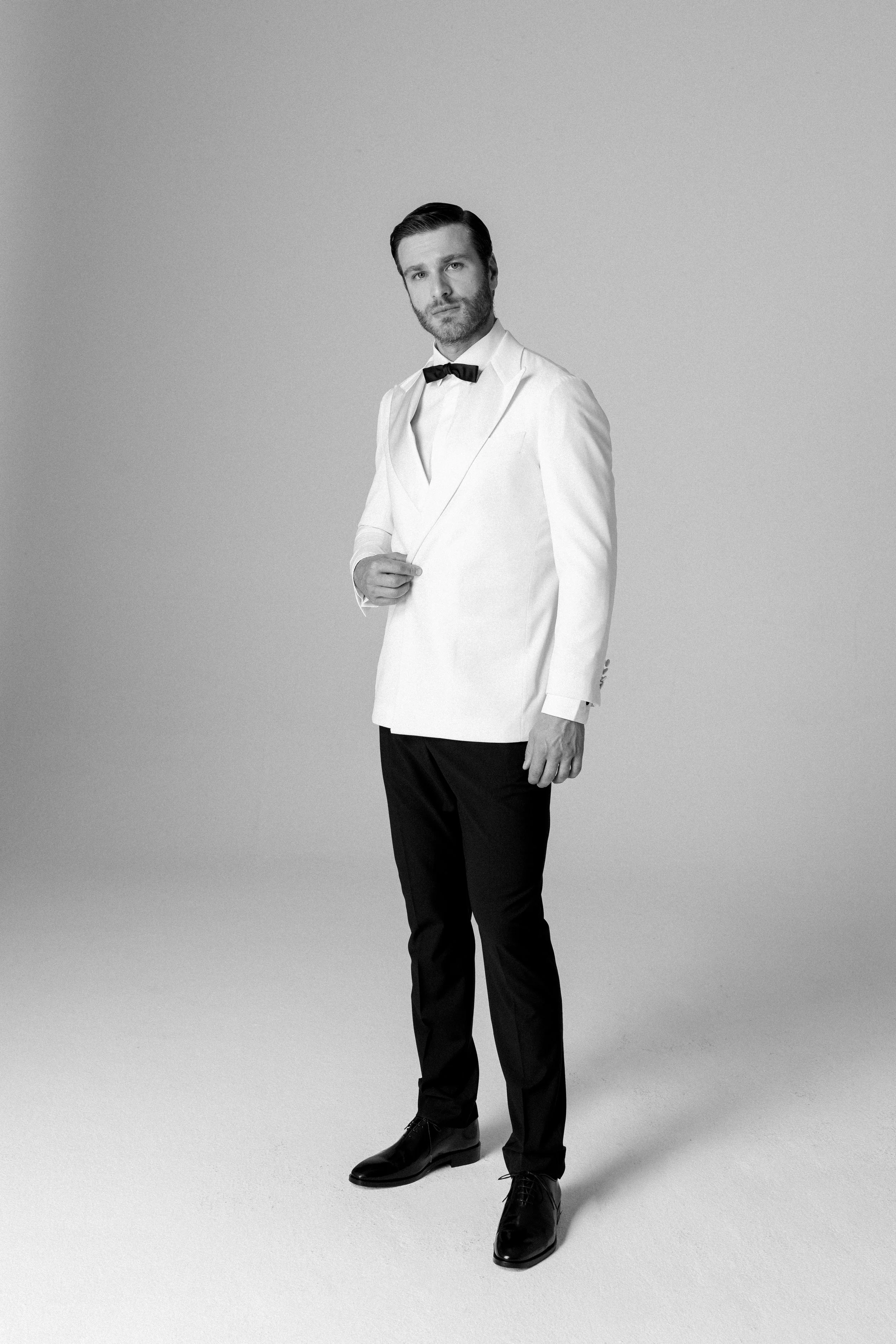 Uomo vestito con smoking bianco e pantaloni neri, indossa un bowler, in piedi contro uno sfondo neutro.