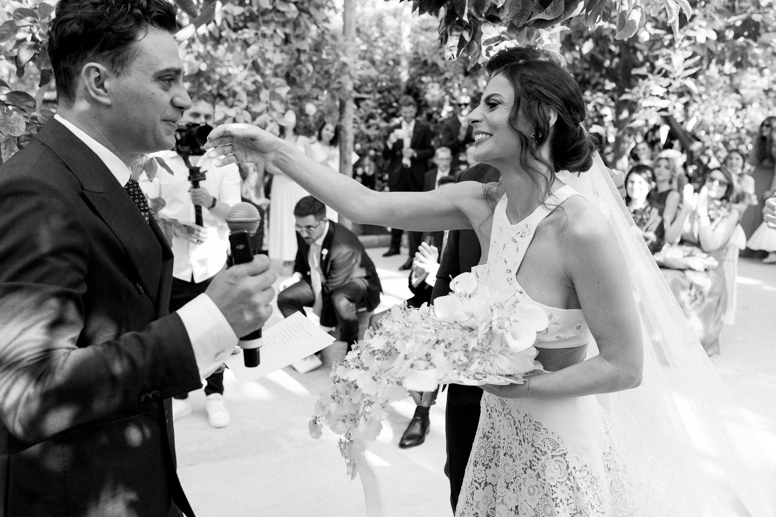 Una coppia si sposa all'aperto, con la sposa che sorride e si avvicina allo sposo, che tiene un microfono. La sposa indossa un abito da sposa in pizzo e tiene un bouquet. Ci sono molti invitati seduti e in piedi che guardano, alcuni sorridono e altri