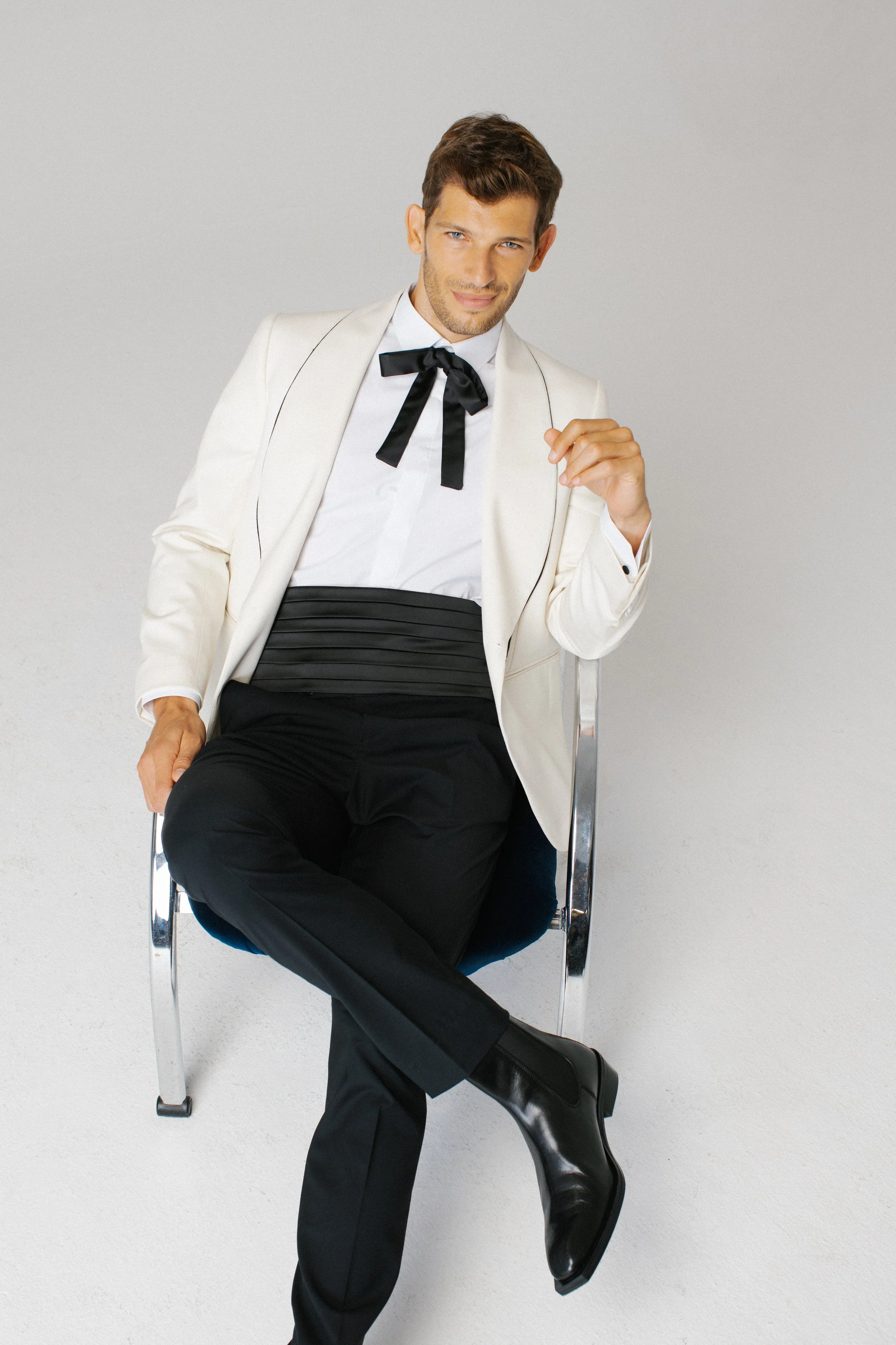 Uomo seduto su una sedia, indossa un abito elegante bianco con pantaloni neri, camicia bianca e cravatta nera, con un sorriso e postura sicura.