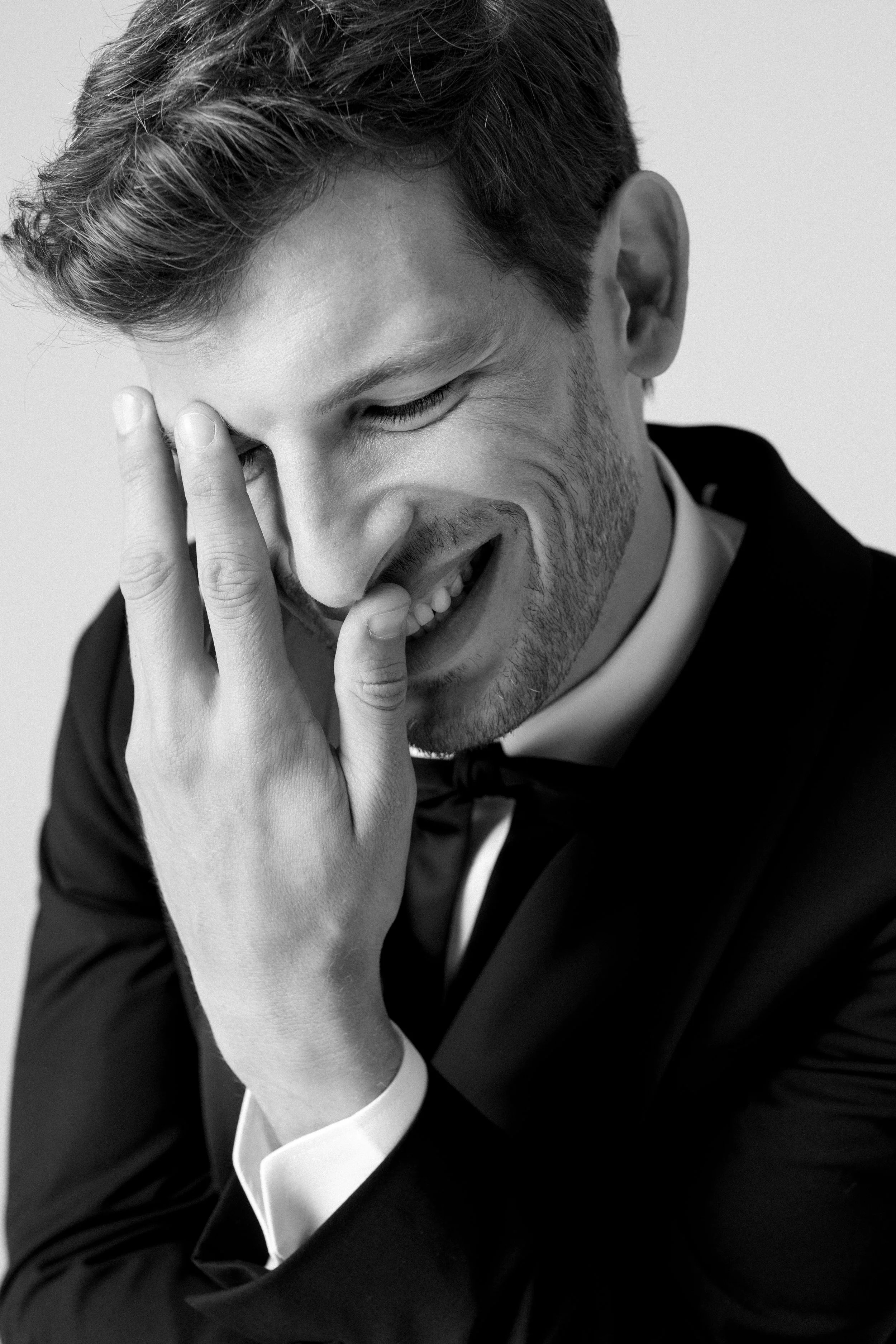 Uomo in smoking nero, con capelli castani, che sorride scherzosamente mentre si tocca il volto con la mano, in bianco e nero.