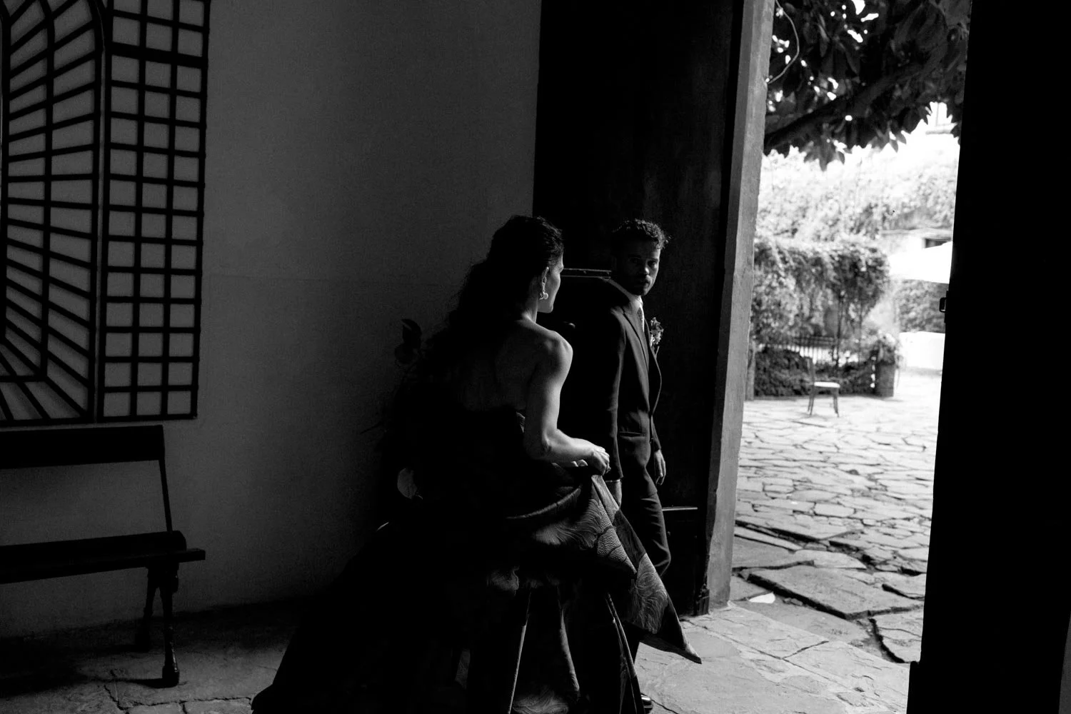 Sposi in un ambiente interno, che si preparano per uscire, guardando verso una porta aperta che mostra un cortile esterno.