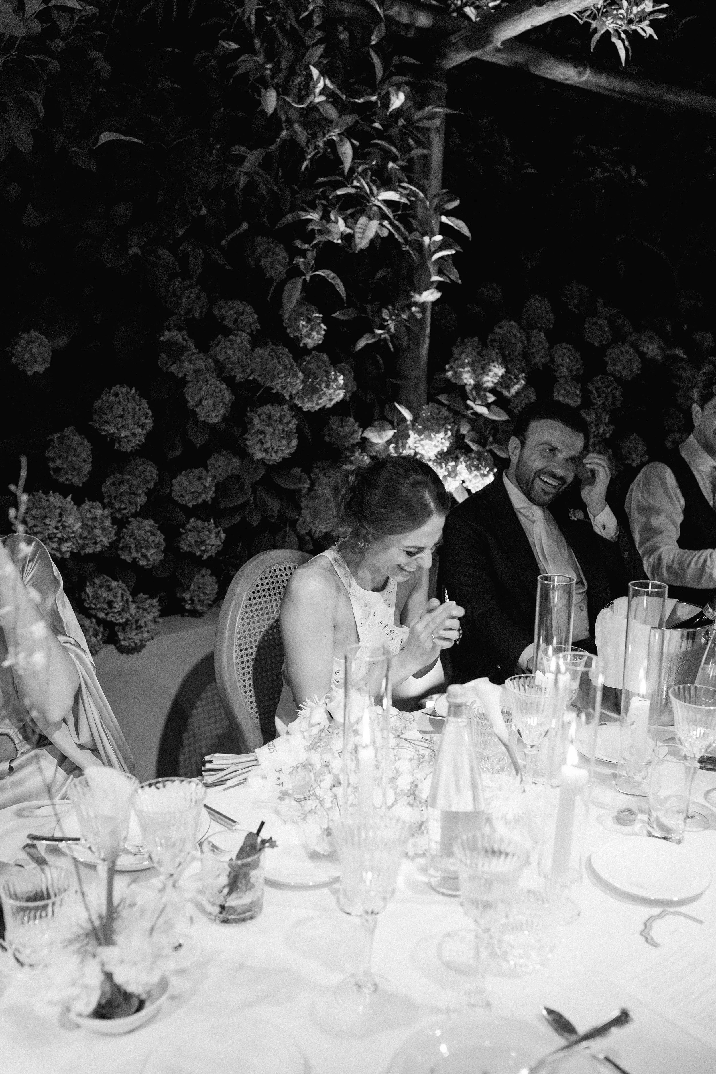 Un gruppo di persone sedute a un tavolo durante un evento, probabilmente un matrimonio, ridendo e sorridendo, con cibo e bicchieri decorativi sulla tavola, e un impianto di fiori sullo sfondo.