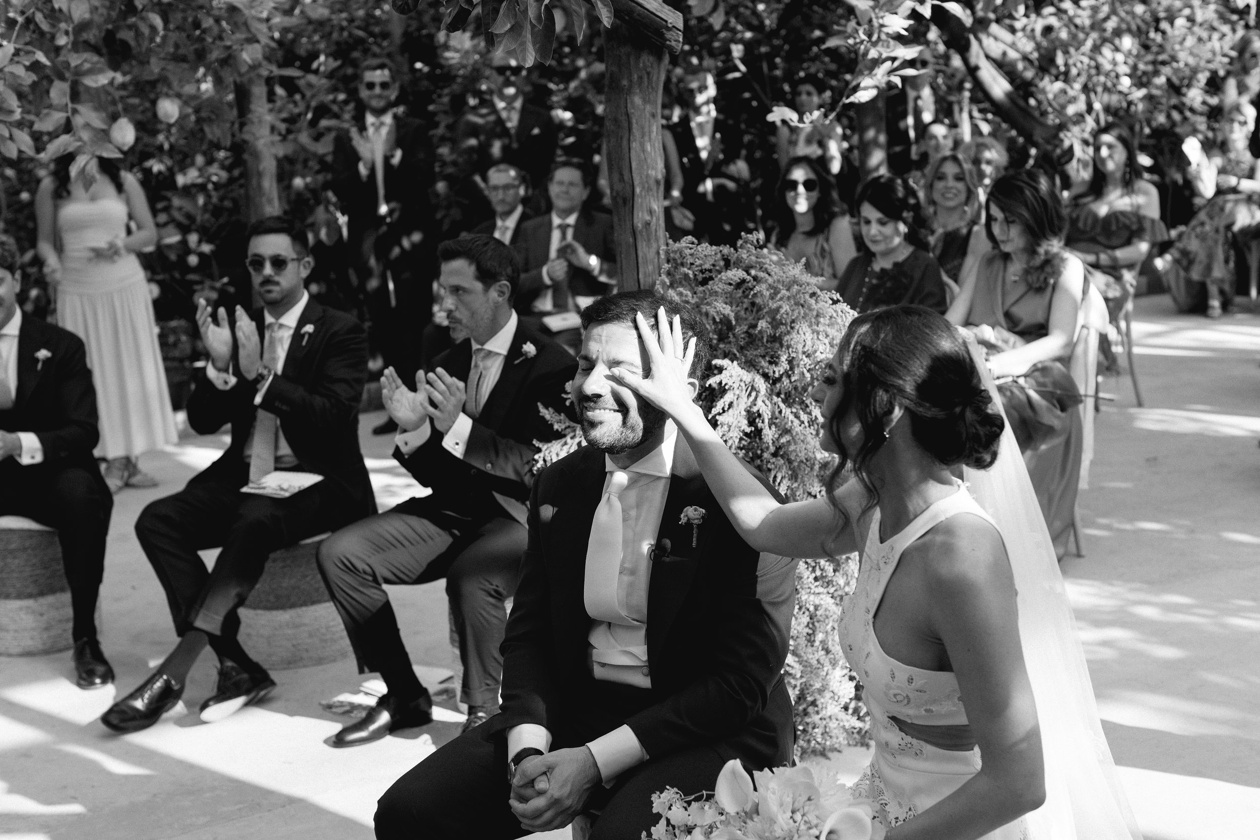 Coppia di sposi in cerimonia di matrimonio, con spettatori seduti e in piedi, decorazioni floreali e ambientazione all'aperto.