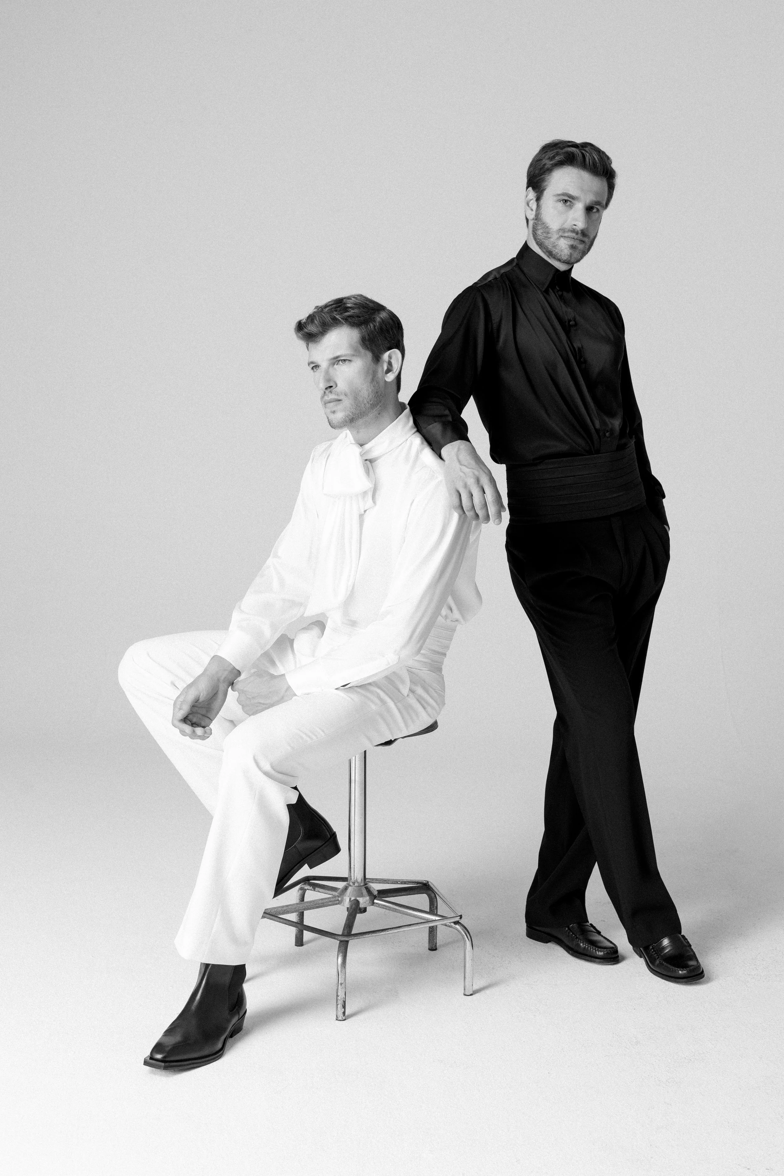 Due uomini vestiti in abiti eleganti, uno seduto e uno in piedi, in una foto in bianco e nero.