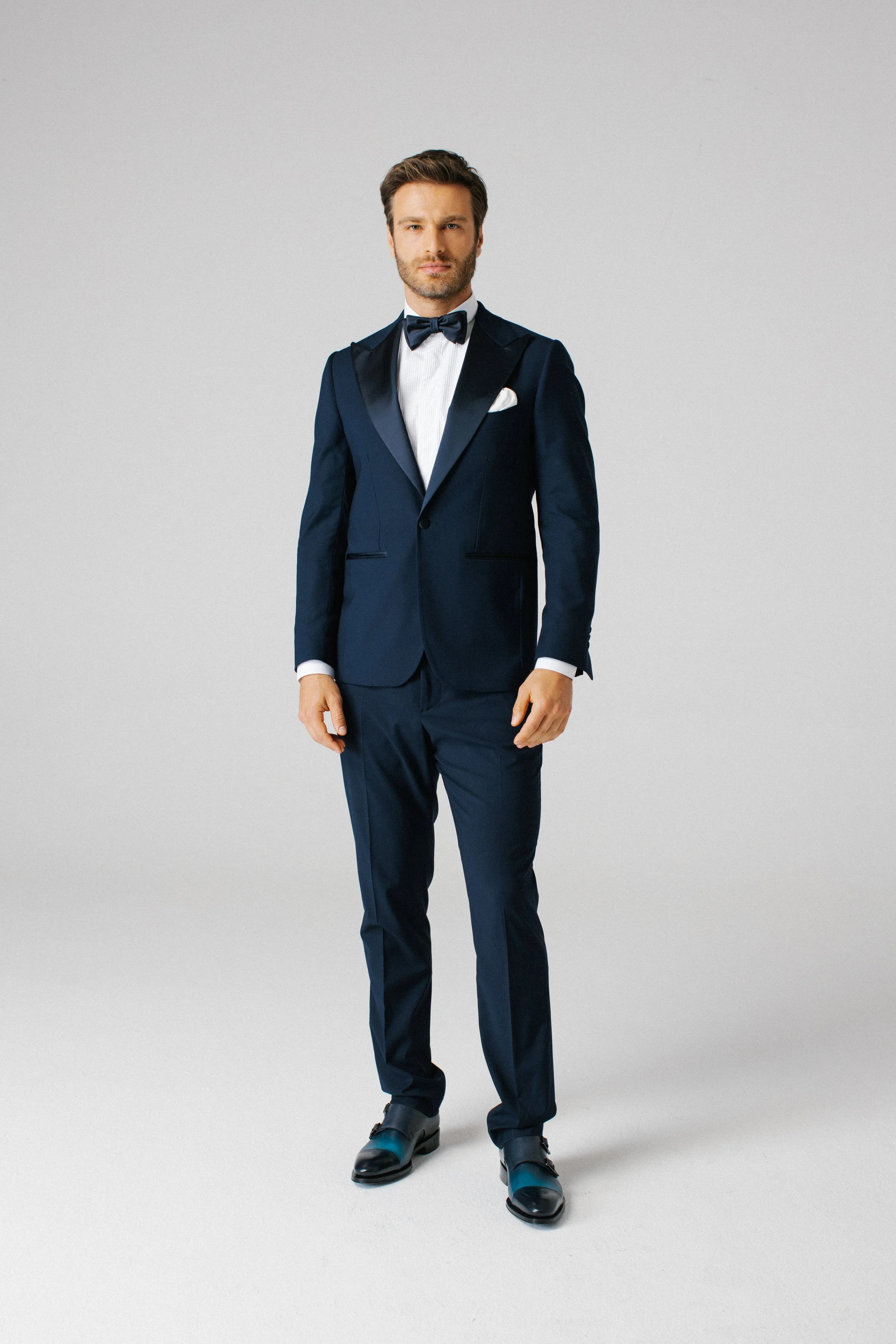 Uomo in abito da sera blu con papillon, posizionato di fronte in uno sfondo grigio chiaro.